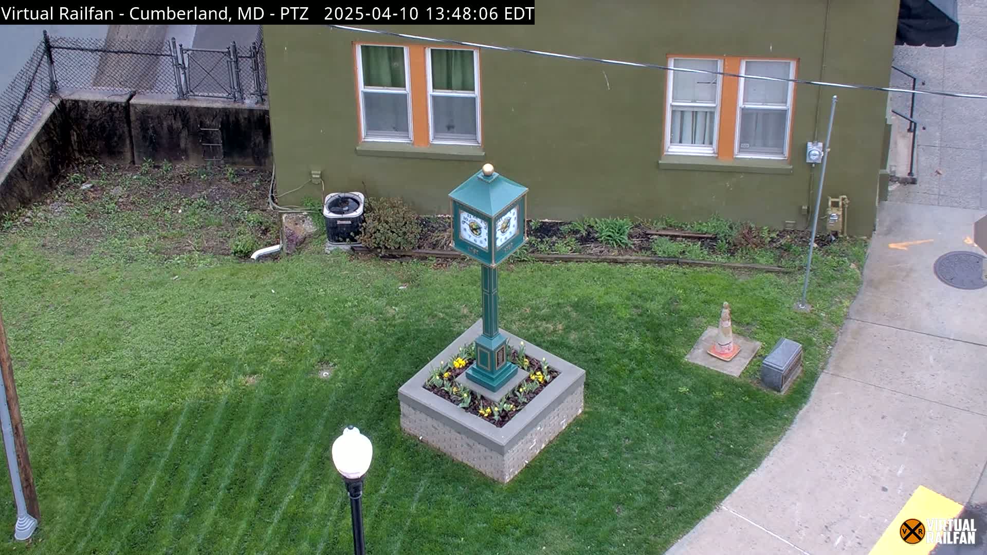 A green clock tower sits in a square flower bed in a grassy area next to a green building on a slightly overcast day.