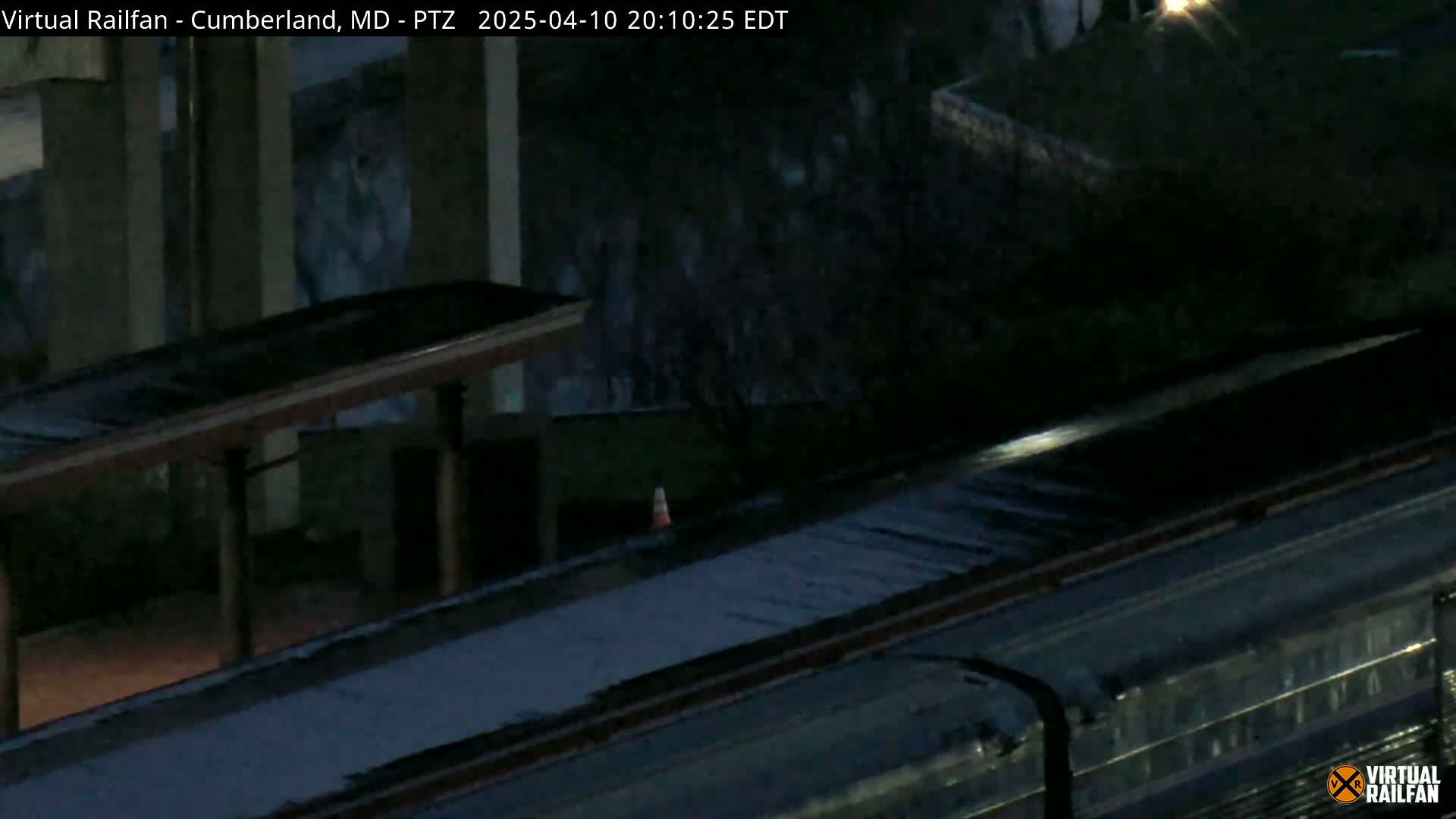 A dark, nighttime image shows a train car parked under a covered shelter next to a building with large murals.
