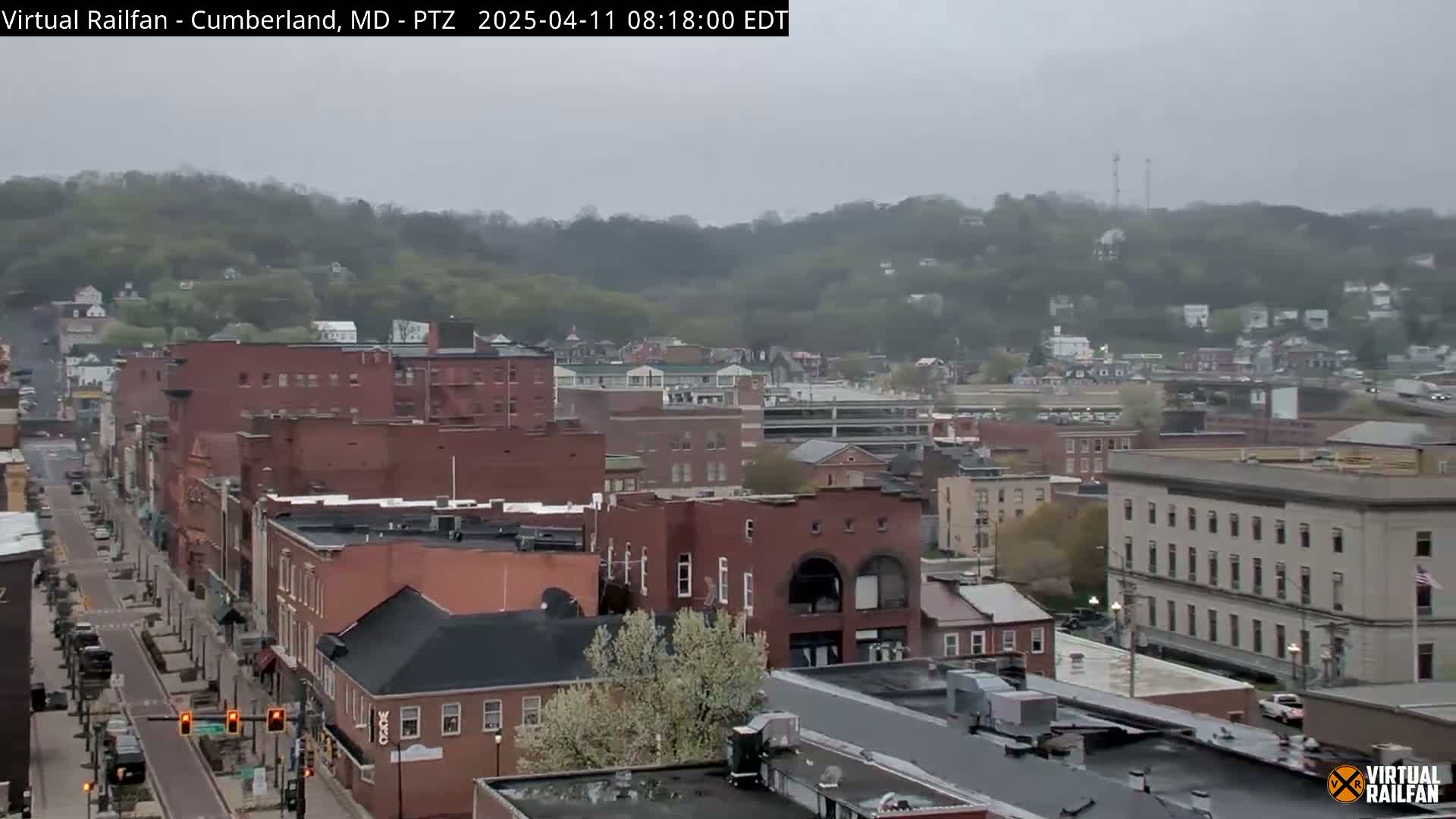 A mostly overcast view of a town with brick buildings and a street running through it, hills covered in trees in the background.