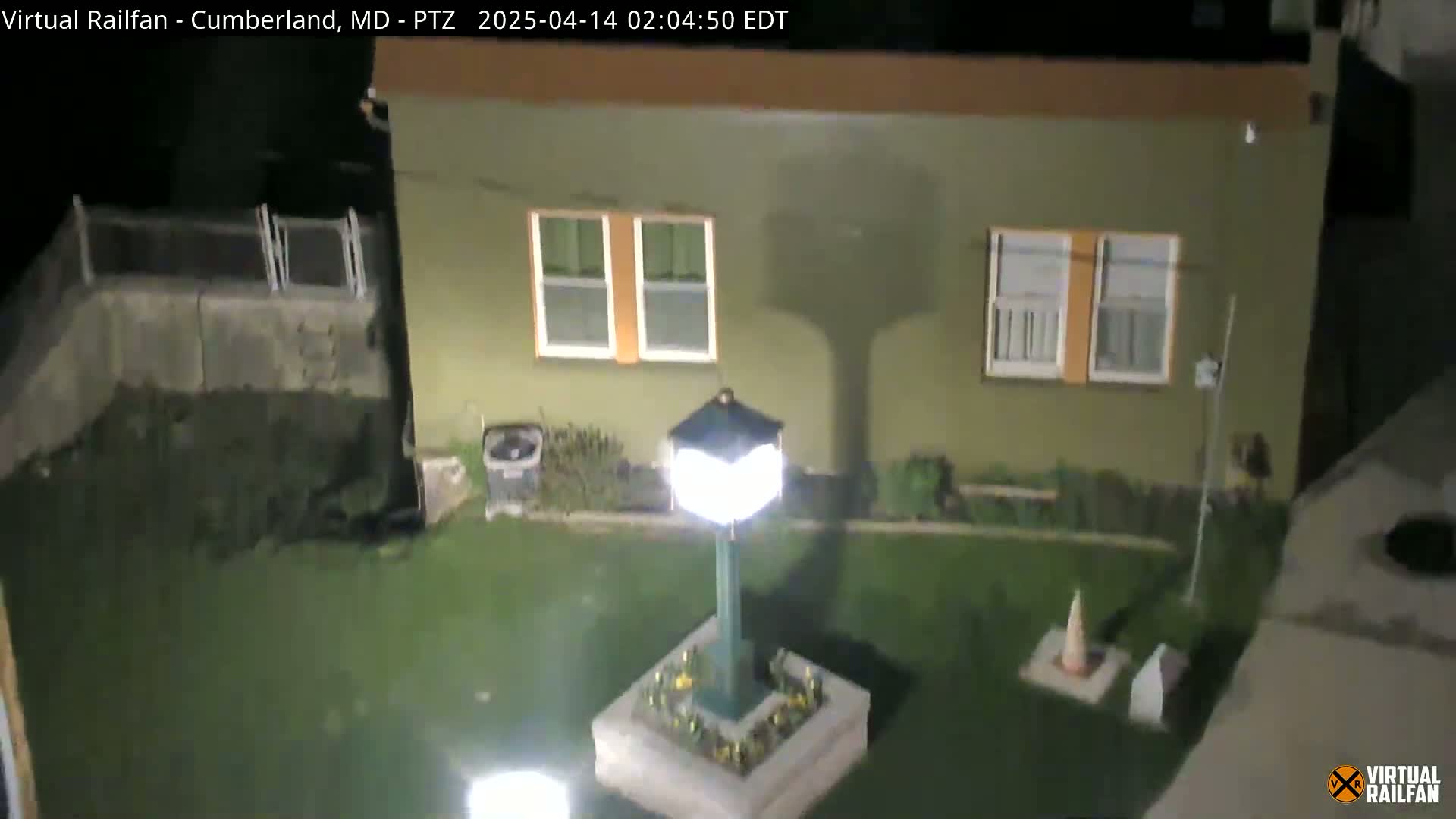 A nighttime view of a small, lit courtyard in front of a tan building, featuring a lamppost in a planter and a small, grassy area.