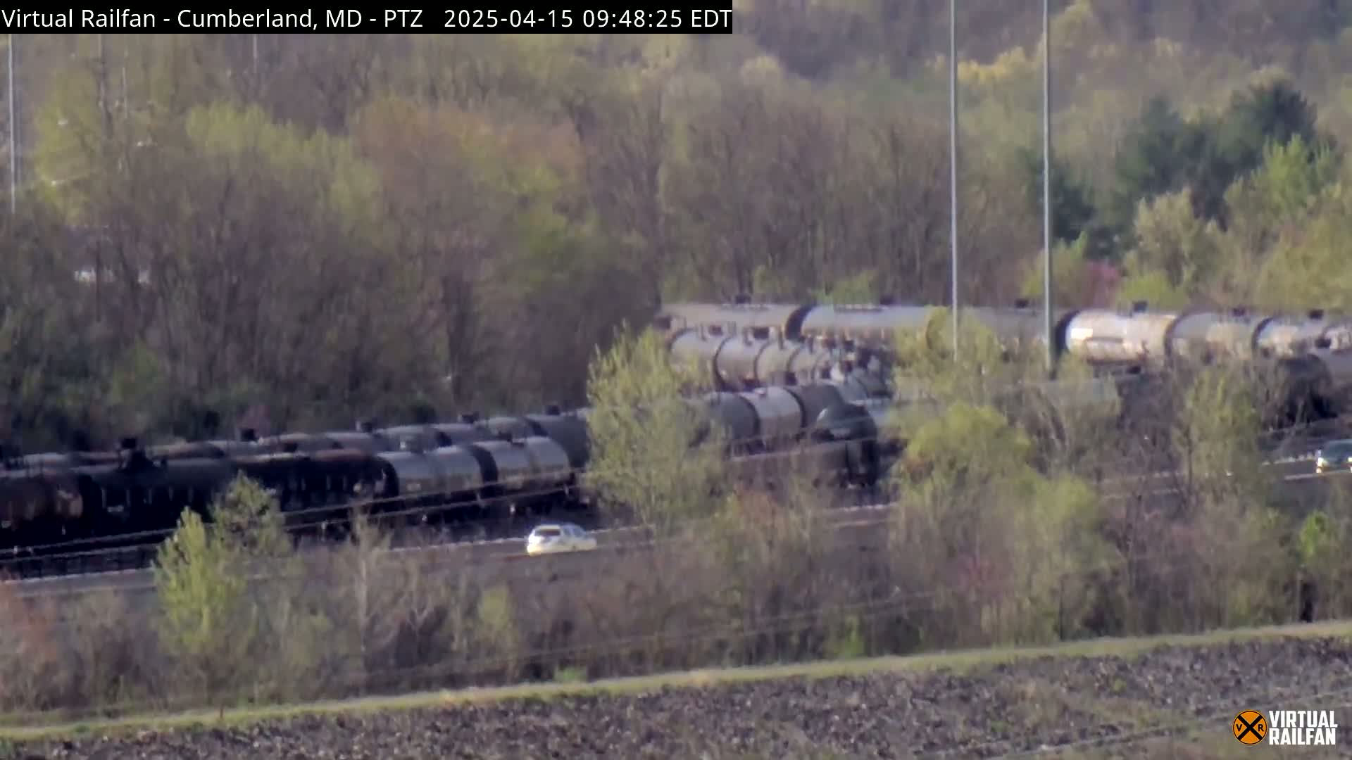 A long freight train of tanker cars sits on a track partially obscured by trees and brush under a mostly sunny sky.