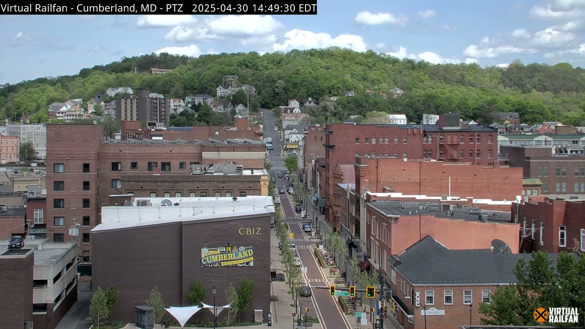 A mostly sunny day reveals a view of a city street lined with brick buildings, ascending towards a tree-covered hill.