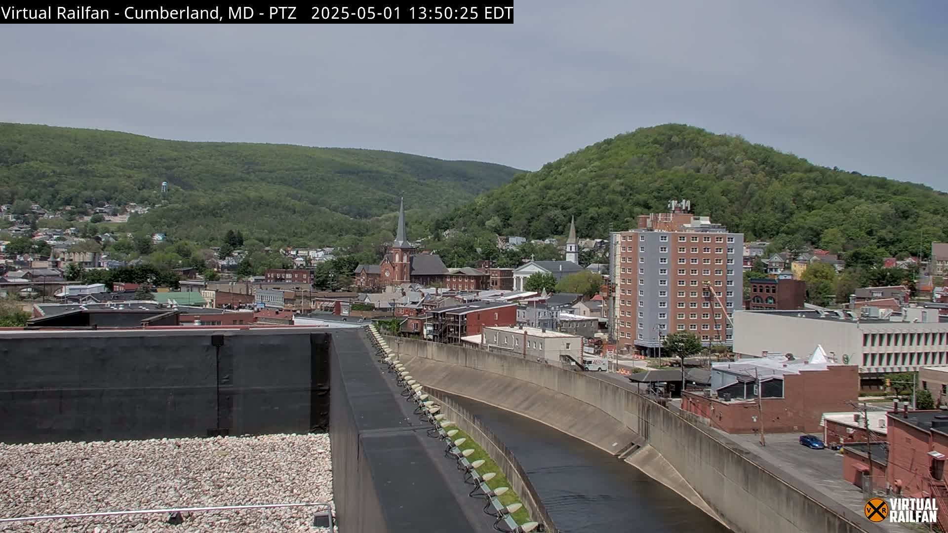 A view of a town nestled beside a river and foothills under a partly cloudy sky.