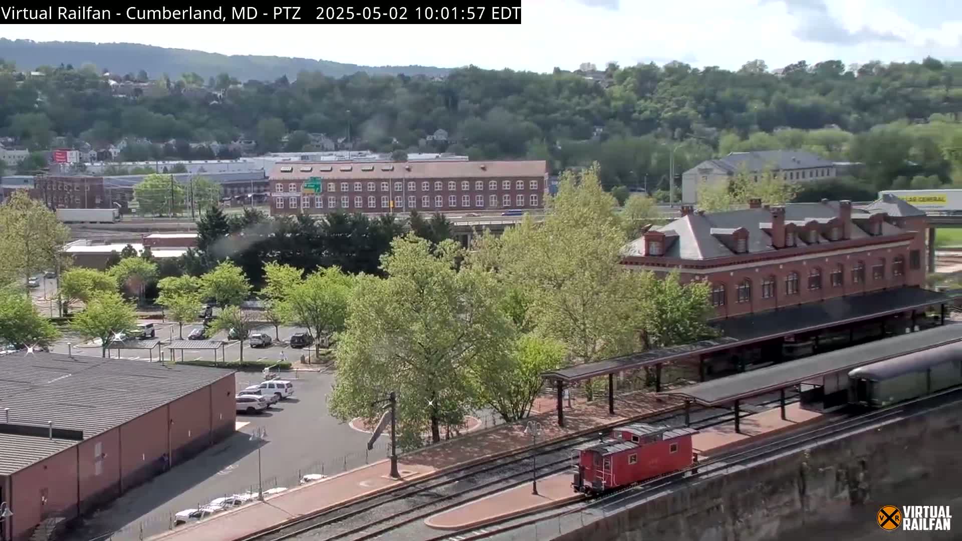 A red caboose and a long black train car sit on train tracks in front of a brick building and parking lot, with green trees and hills in the background on a sunny day.