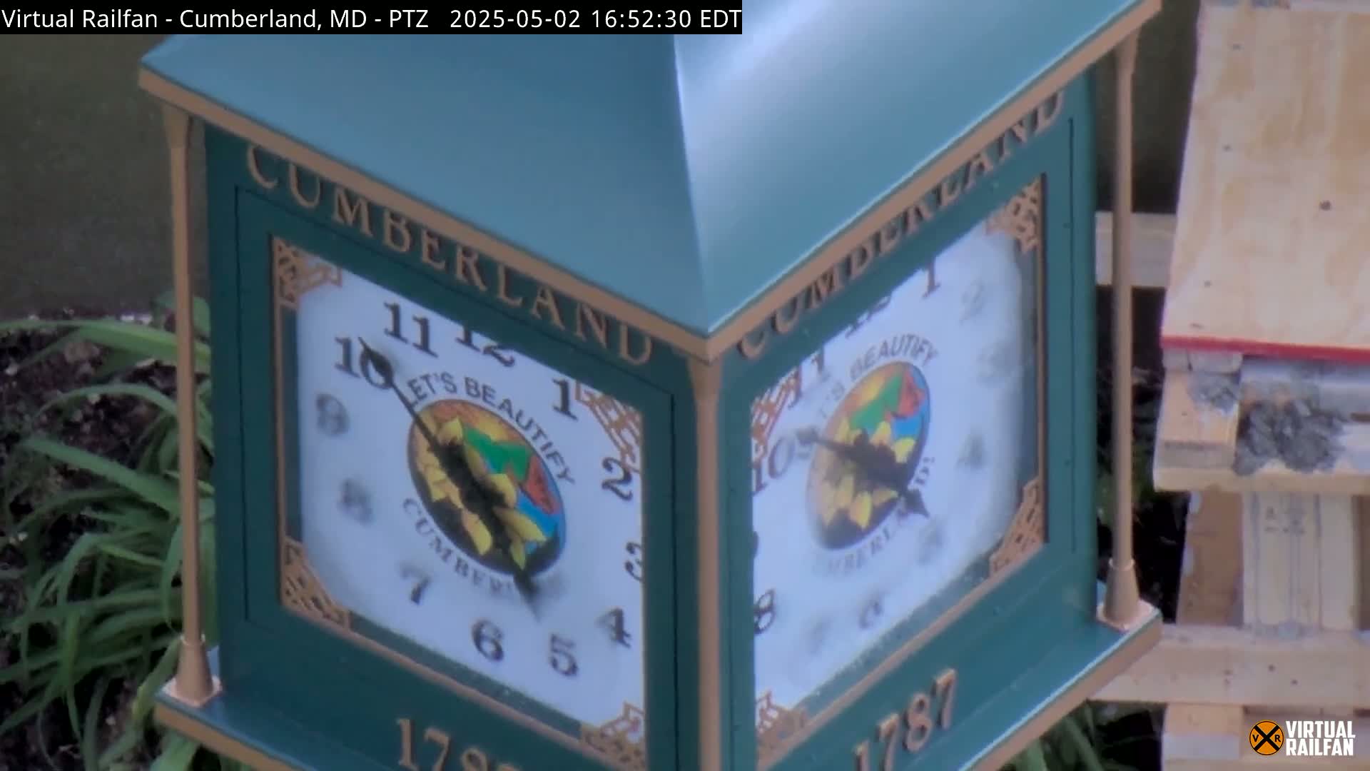 A green, multi-sided clock with "Cumberland" and a colorful logo is partially visible, next to some greenery and construction materials.