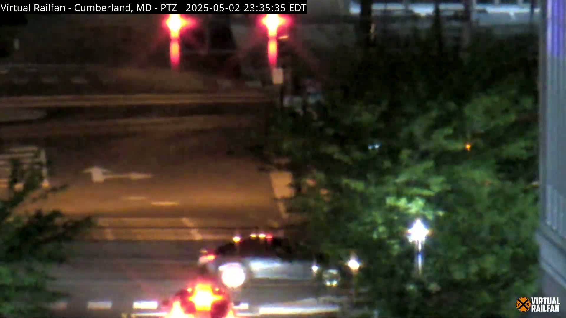 At night, two cars are visible on a street, partially obscured by trees, with red signal lights flashing in the background.