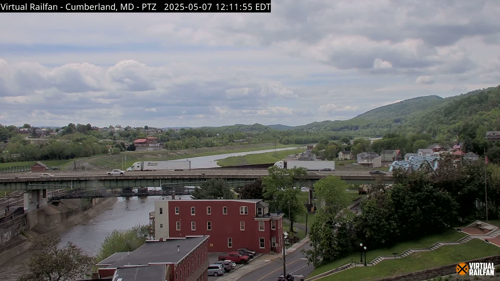 A partly cloudy day reveals a river flowing beneath a bridge, with a town nestled beside it and green hills in the background.