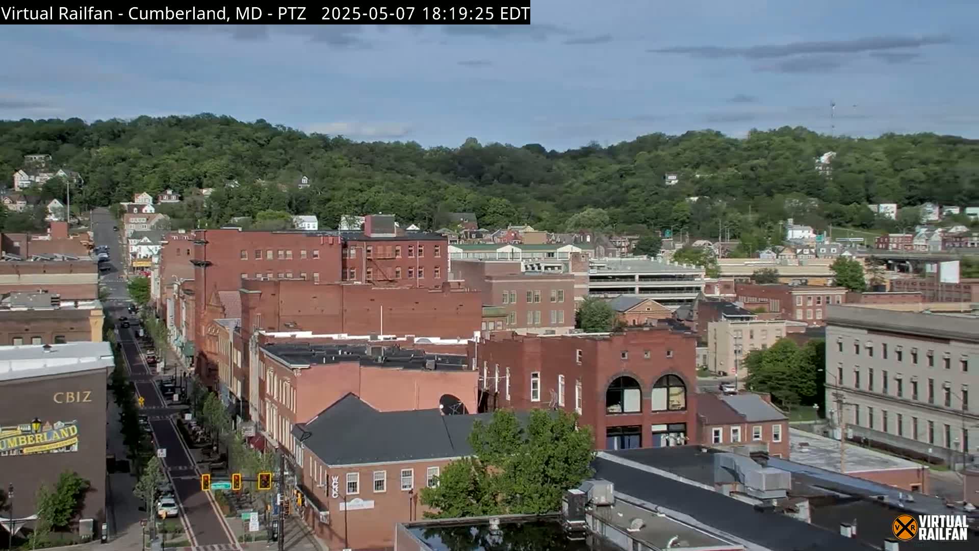 A mostly sunny view of a city street lined with brick buildings, nestled against a green hillside.