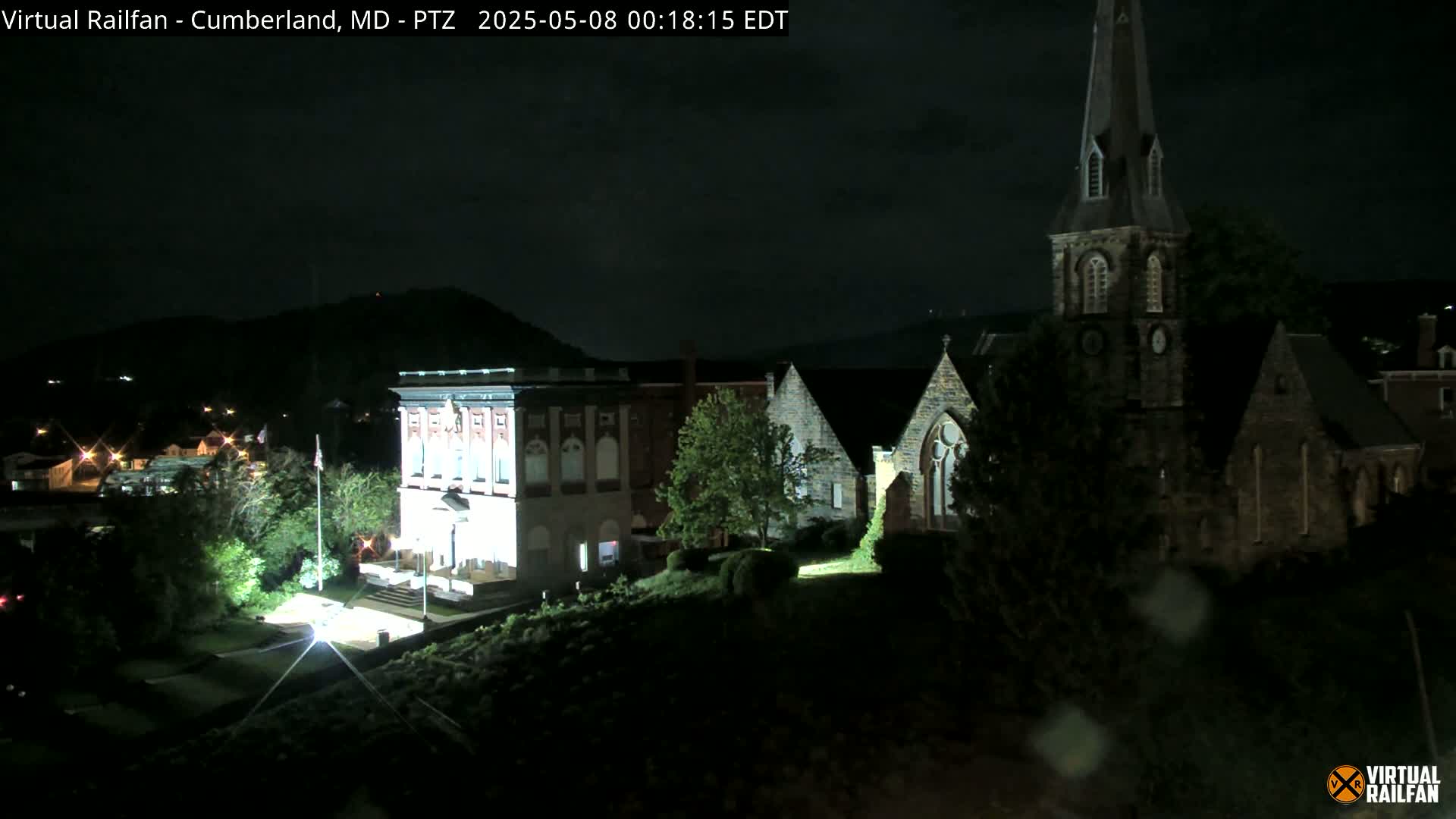 A nighttime view shows a brightly lit brick building and a dark stone church, set against a backdrop of dark hills under a cloudy sky.
