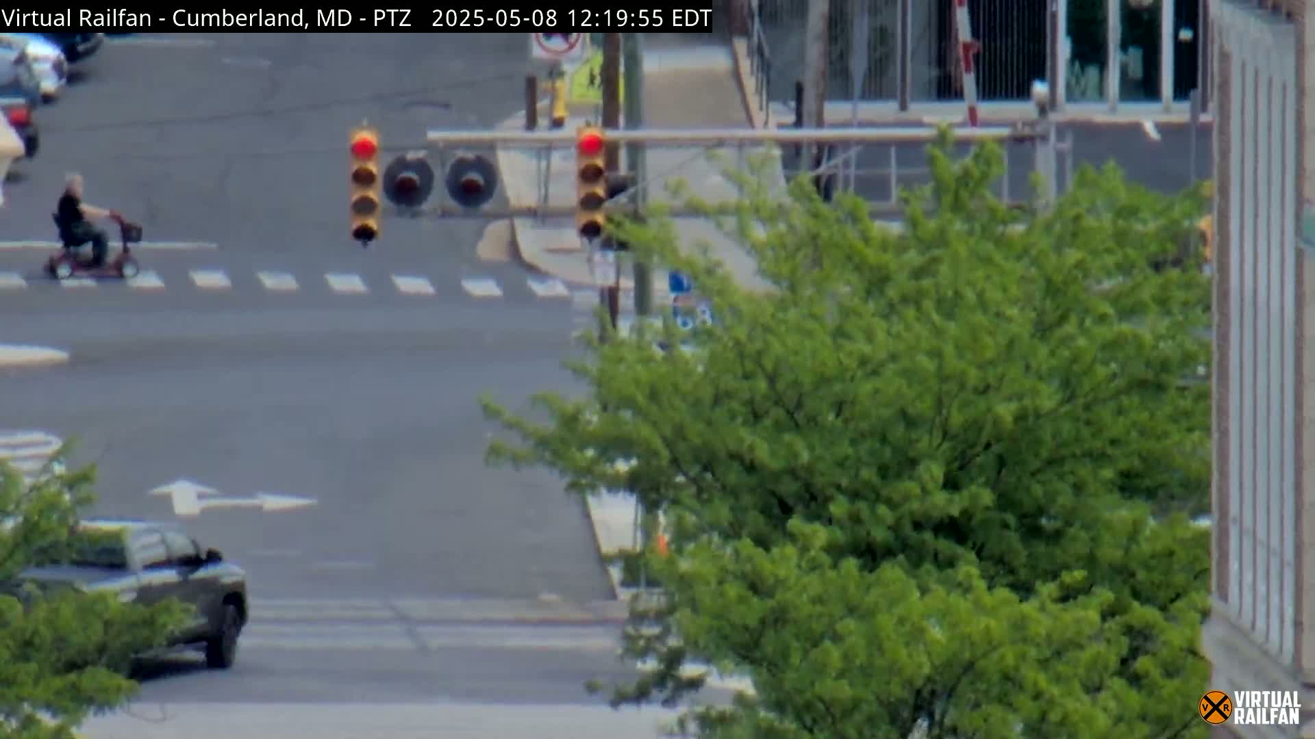 A person on a motorized scooter crosses a street intersection with a dark-colored pickup truck and traffic lights, on a sunny day.