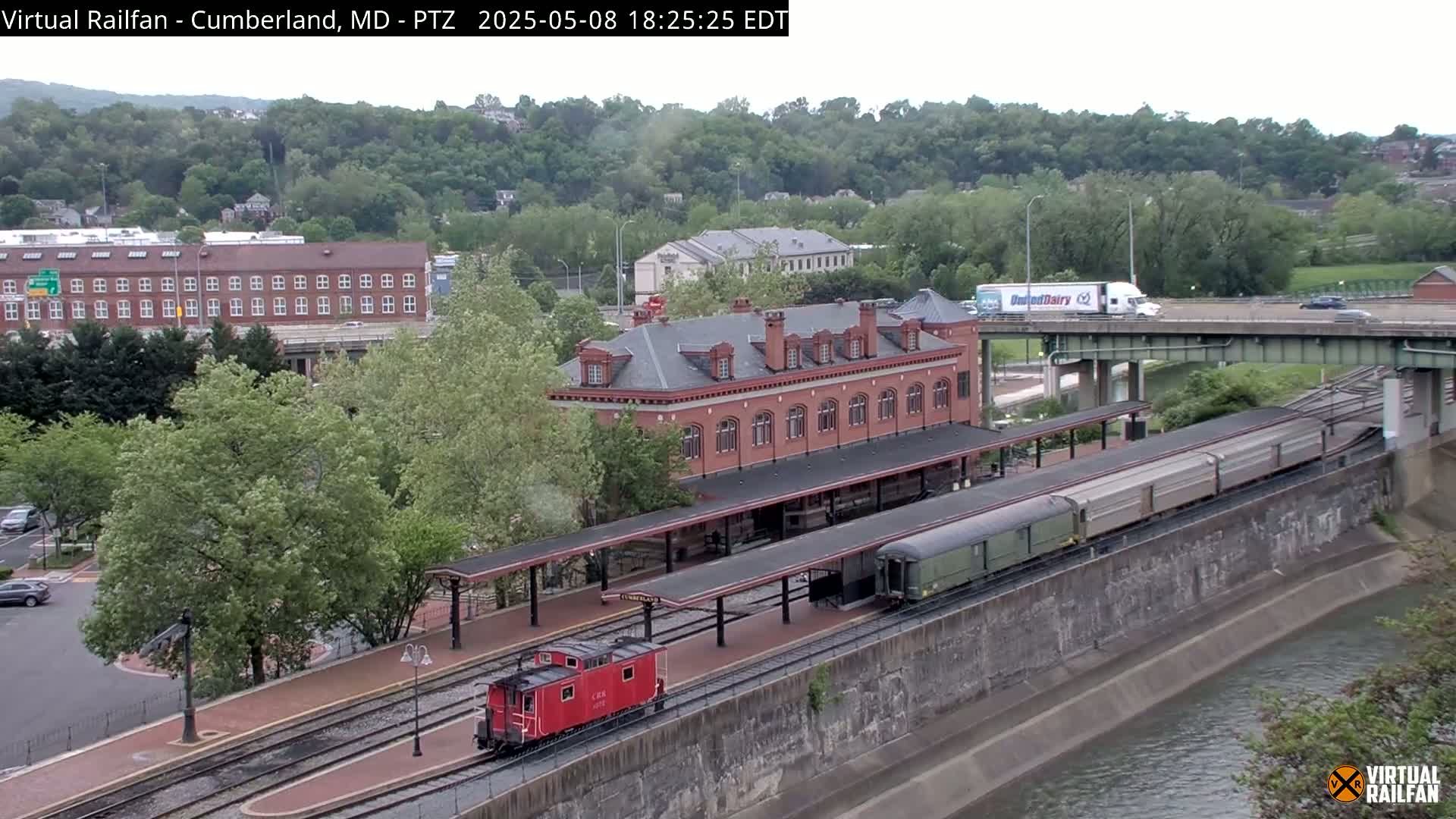 A red caboose and a long train are parked at a brick train station next to a river, under an overcast sky.