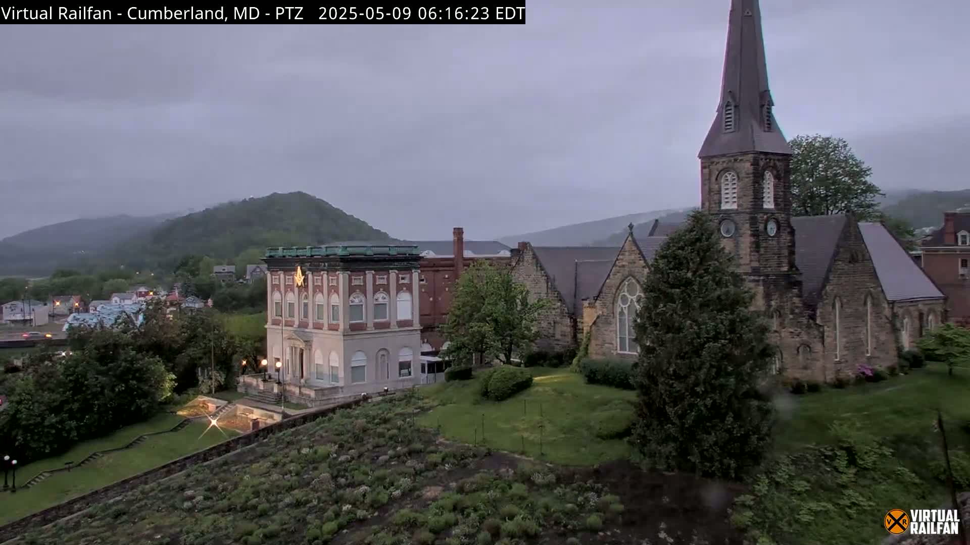 A view of a town nestled in a valley under an overcast sky, featuring a stone church with a tall steeple and a multi-story building.