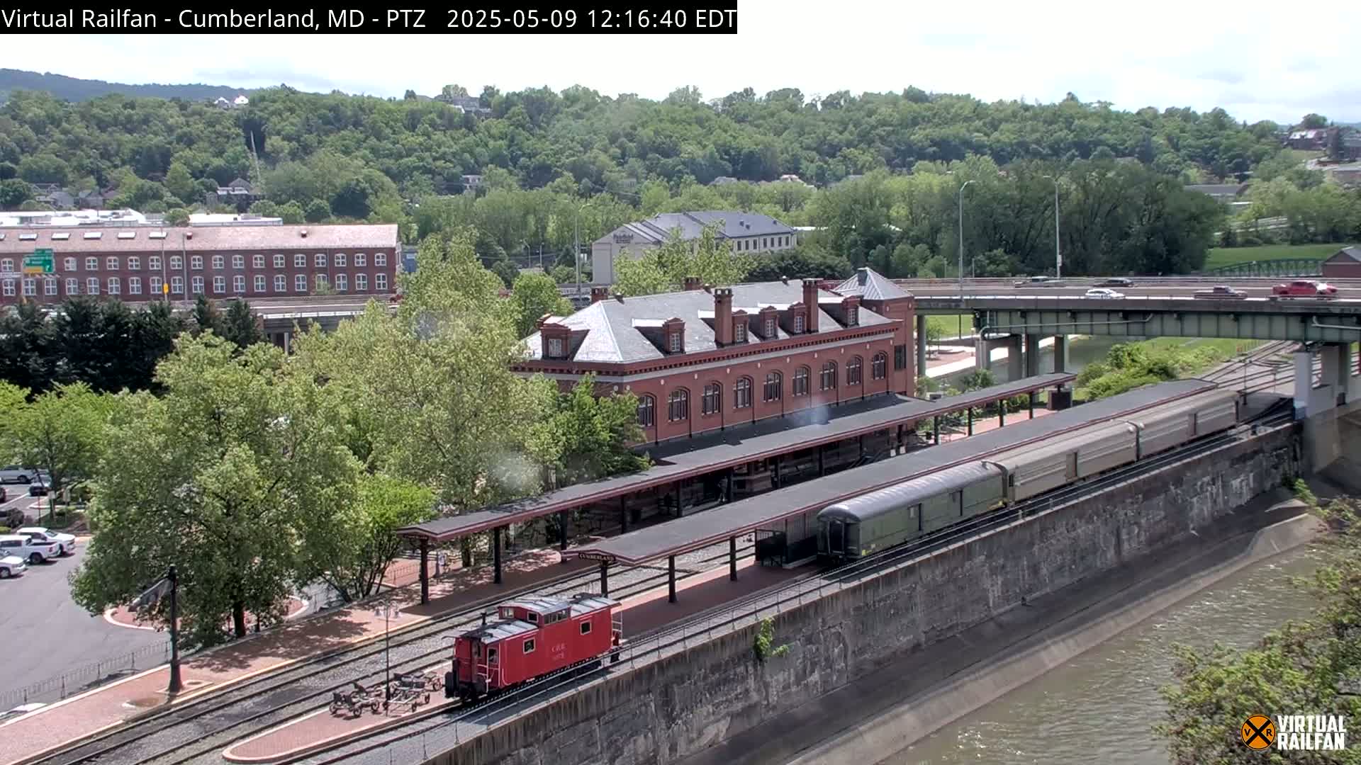 A train sits at a station next to a brick building, with a river and highway visible in the background on a sunny day.