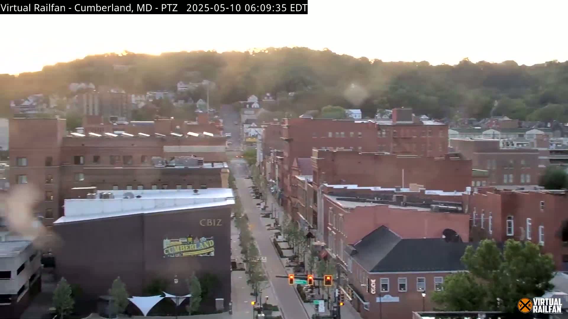A mostly clear sunrise illuminates a long, tree-lined street in a city with mostly brick buildings, hills visible in the background.
