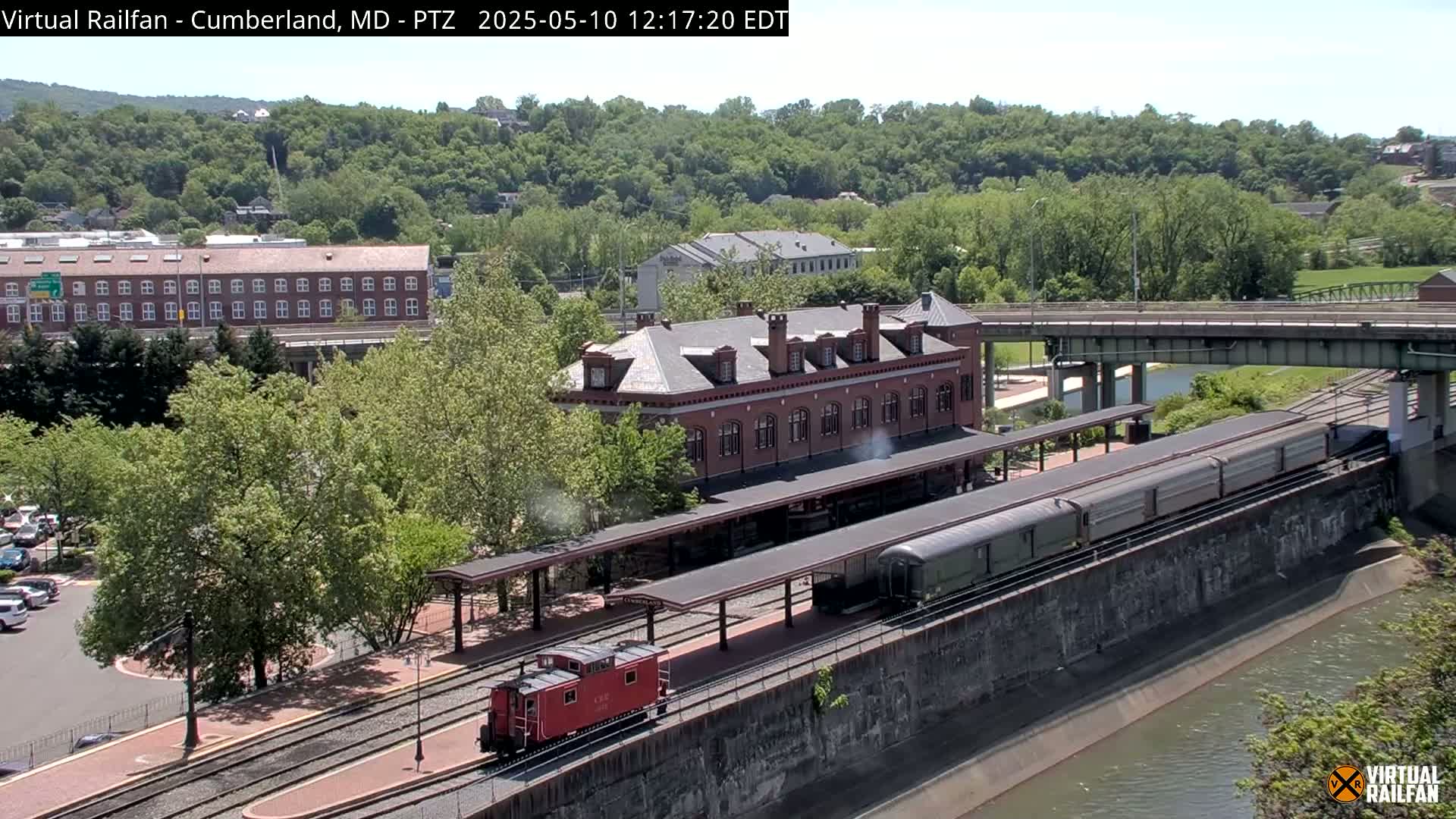 A long passenger train sits at a brick train station next to a river under a sunny sky, with lush green trees and buildings in the background.