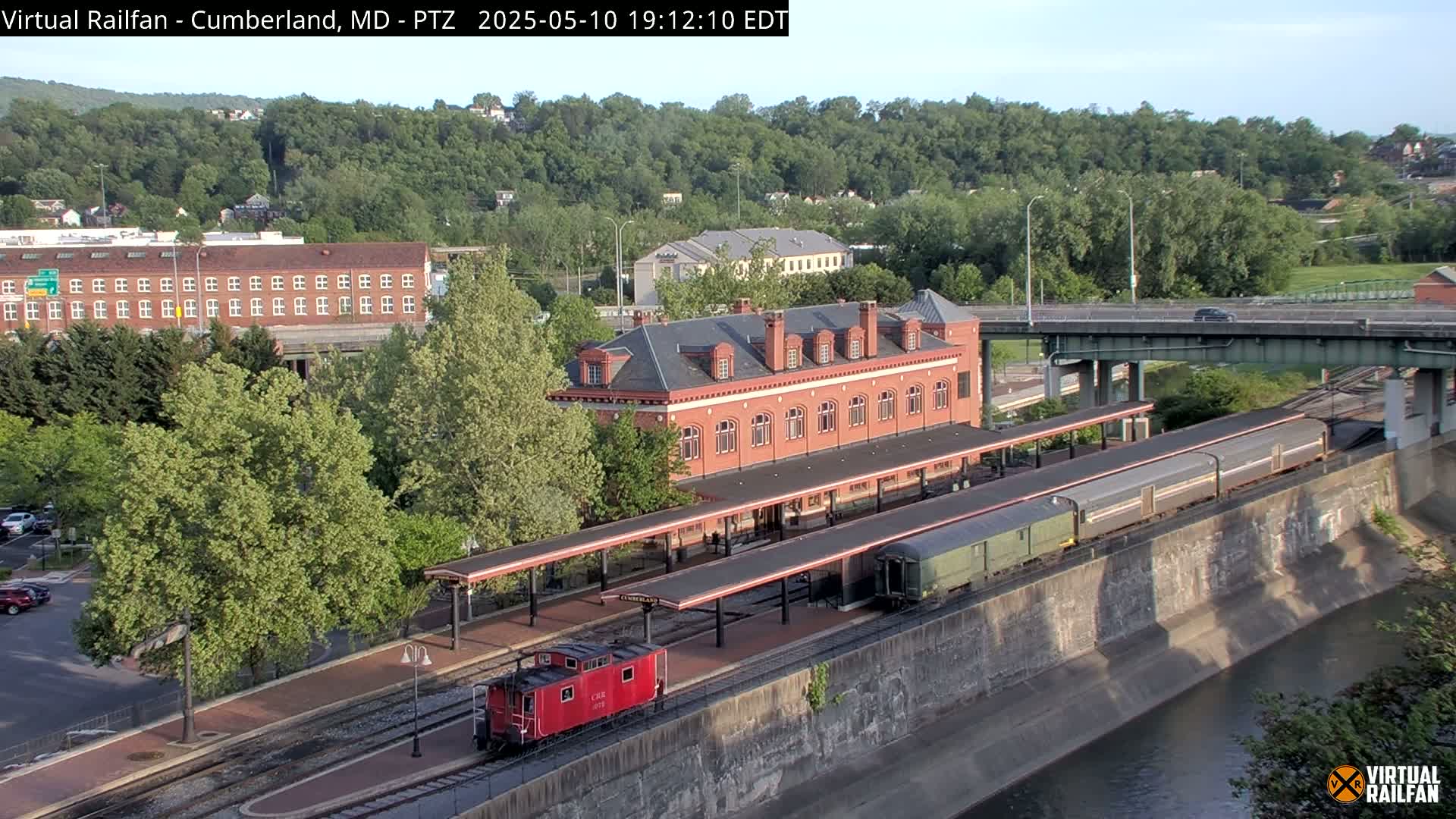 A red caboose and a long train sit at a brick train station next to a river under a mostly sunny sky.