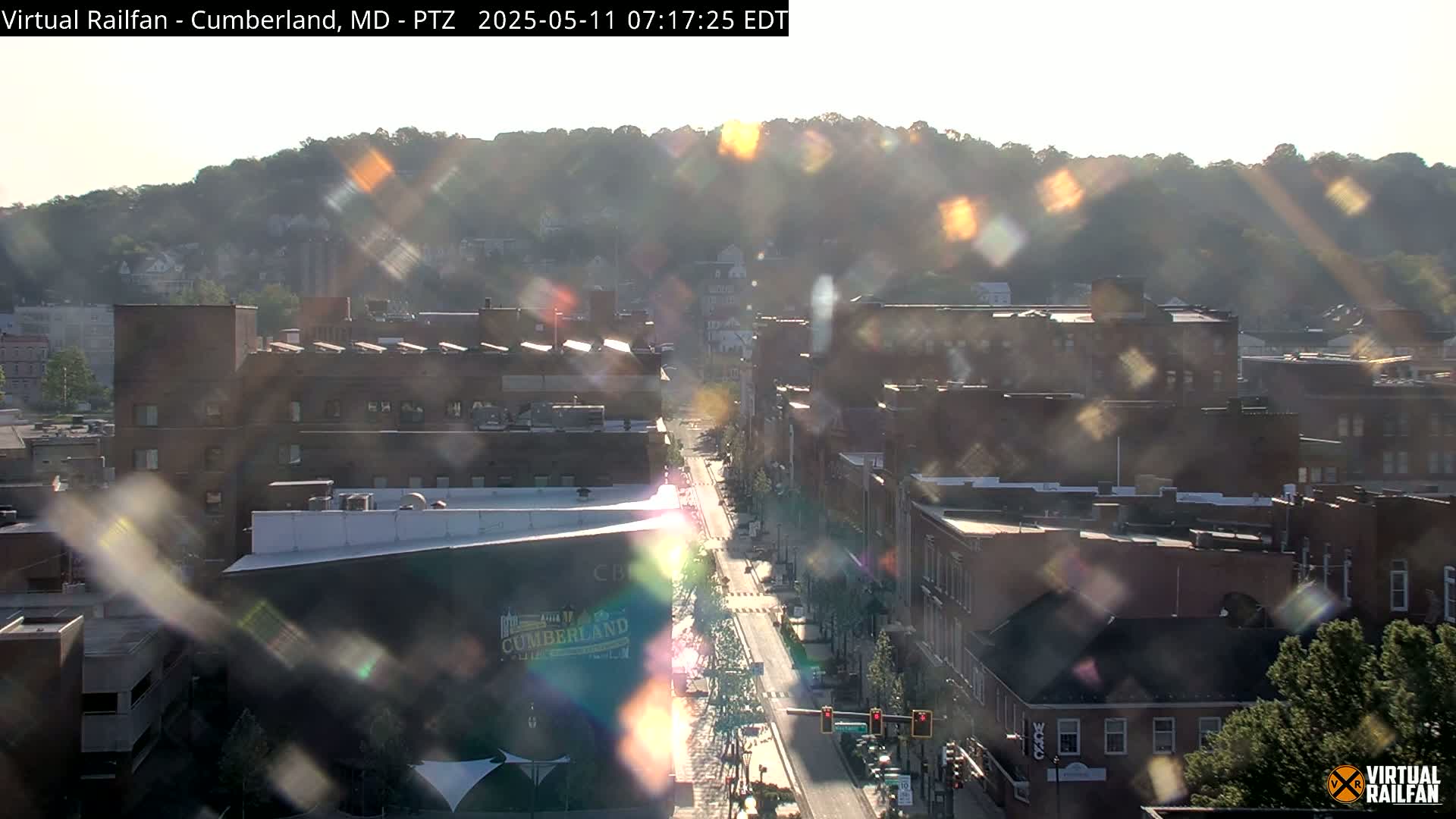A hazy, sunlit view of a city street lined with brick buildings, nestled at the base of a tree-covered hill.