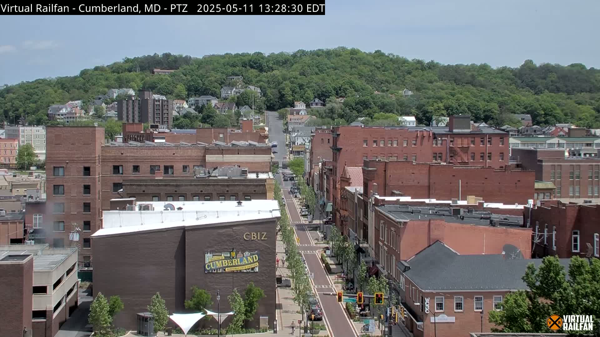 A mostly sunny view of a city street lined with brick buildings,  with a tree-covered hill in the background.