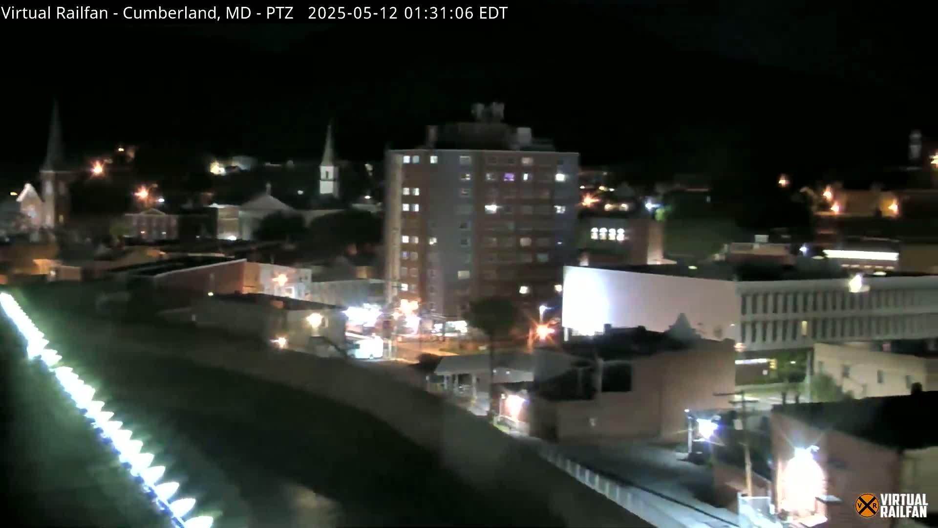 A nighttime view of a town shows a multi-story building surrounded by smaller buildings, lit streets, and a line of lights along a structure.
