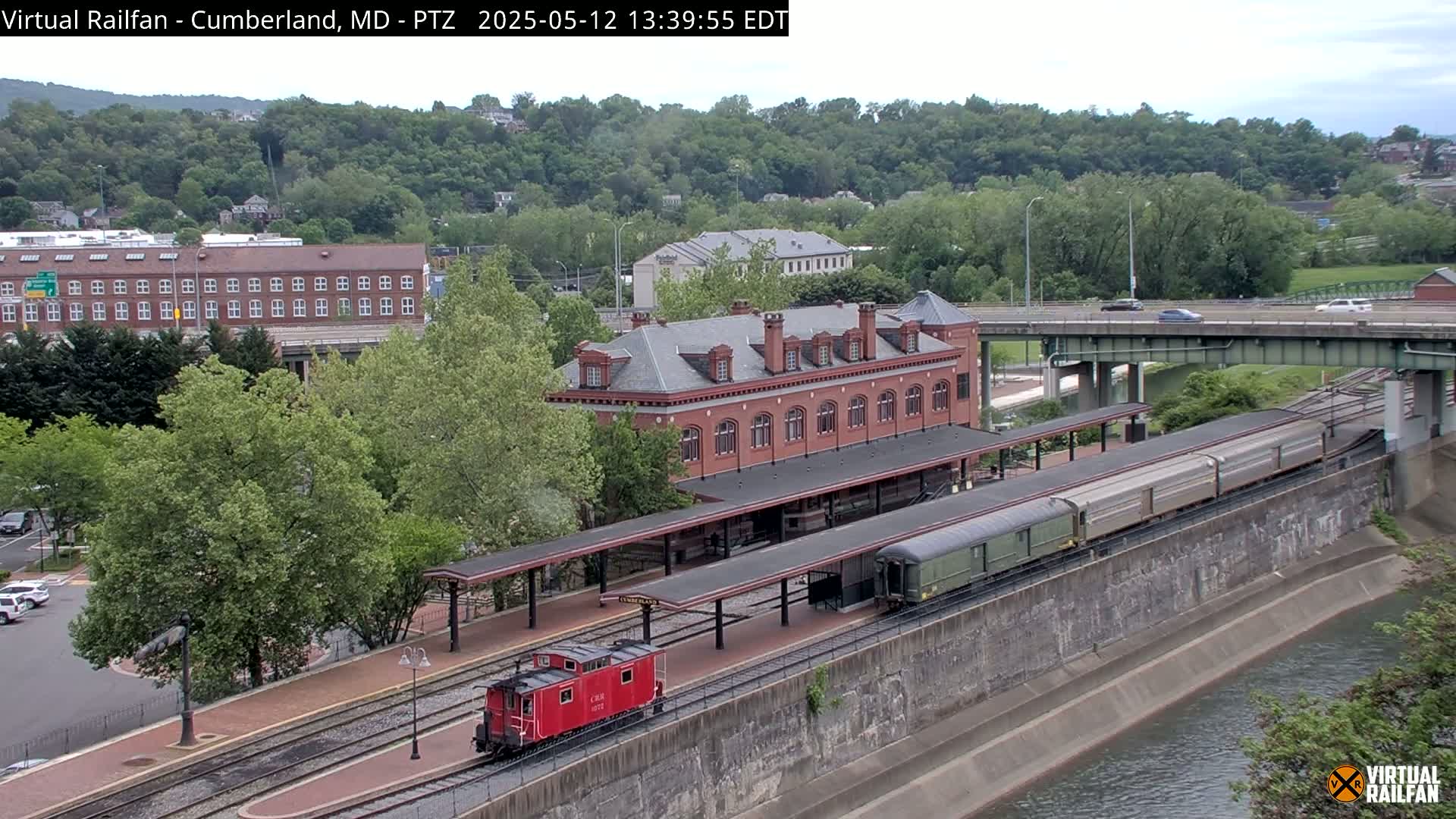 A red caboose and a long train are parked at a brick train station beside a river under a partly cloudy sky.