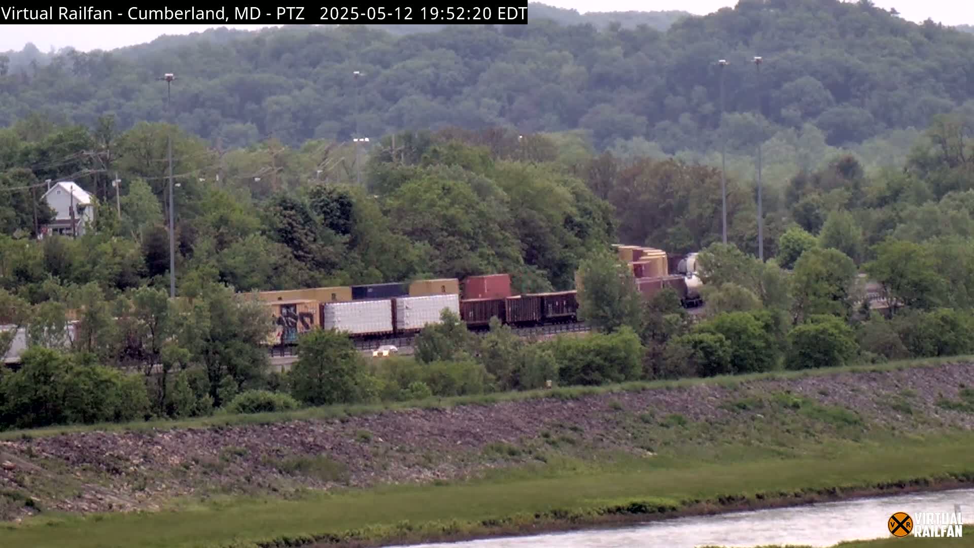 A freight train moves through a lush green landscape beside a river under an overcast sky.