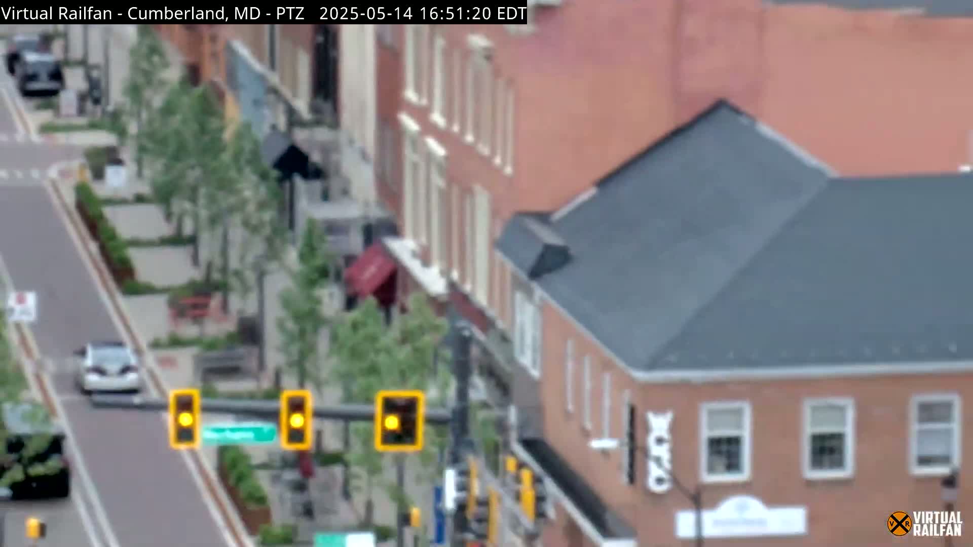 A slightly overcast view of a street scene shows a car driving past traffic signals near brick buildings and landscaped areas.