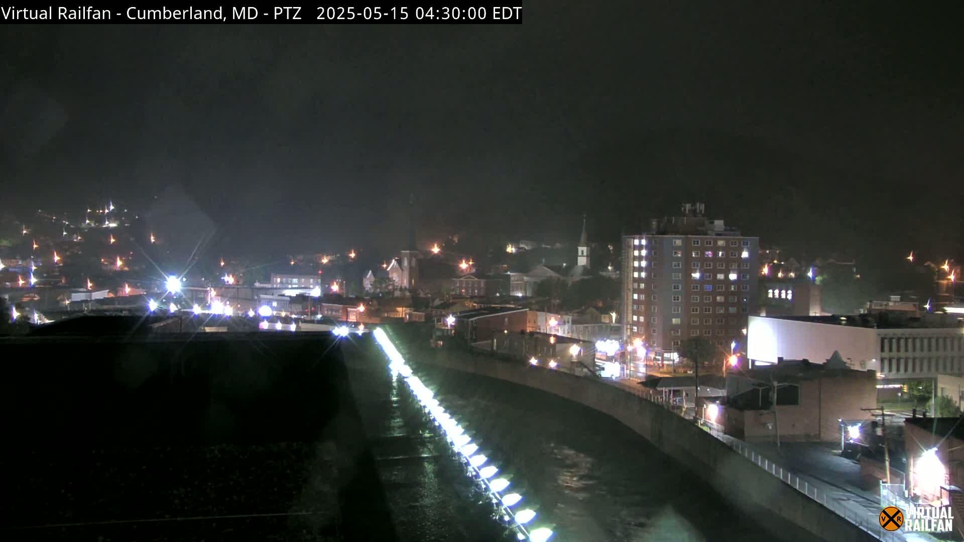 A nighttime view of a rain-slicked town beside a river, illuminated by streetlights and building lights.