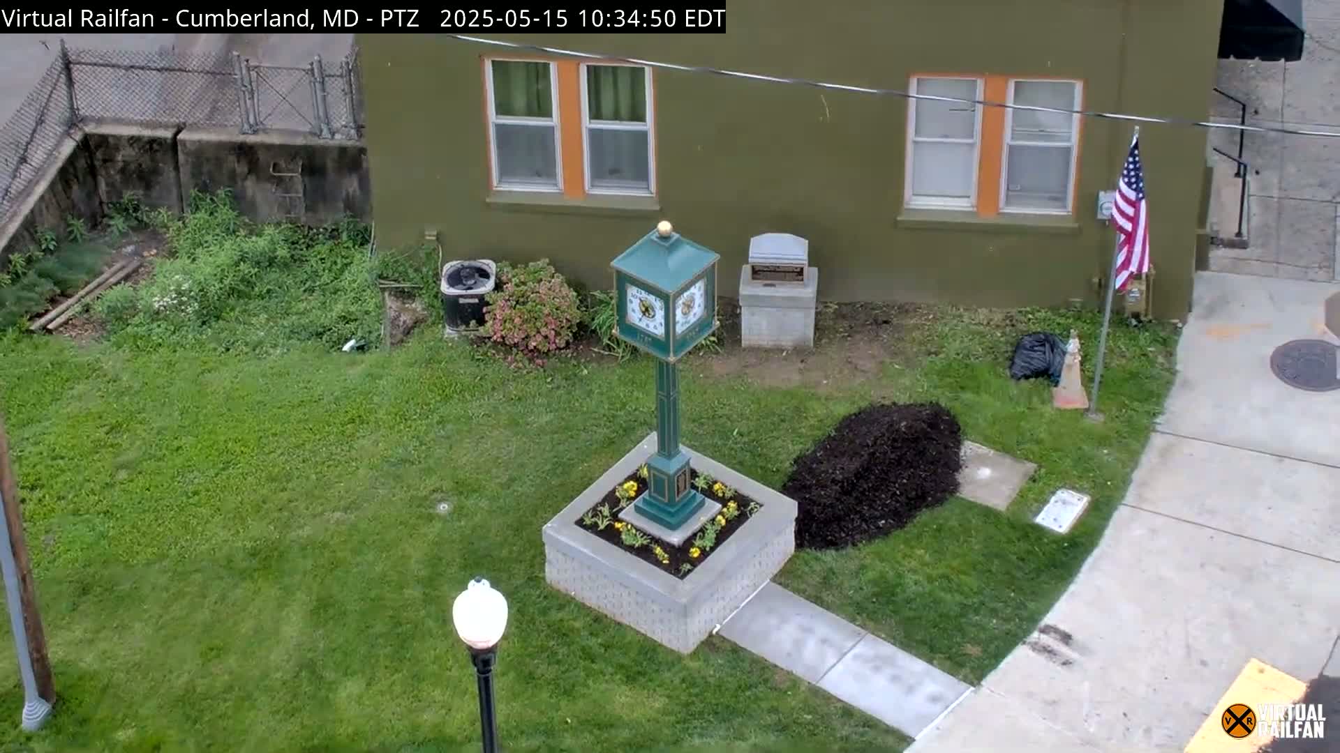 A green clock stands in a square planter surrounded by grass and a recently-mulched area, next to a green building with an American flag and a pile of mulch.
