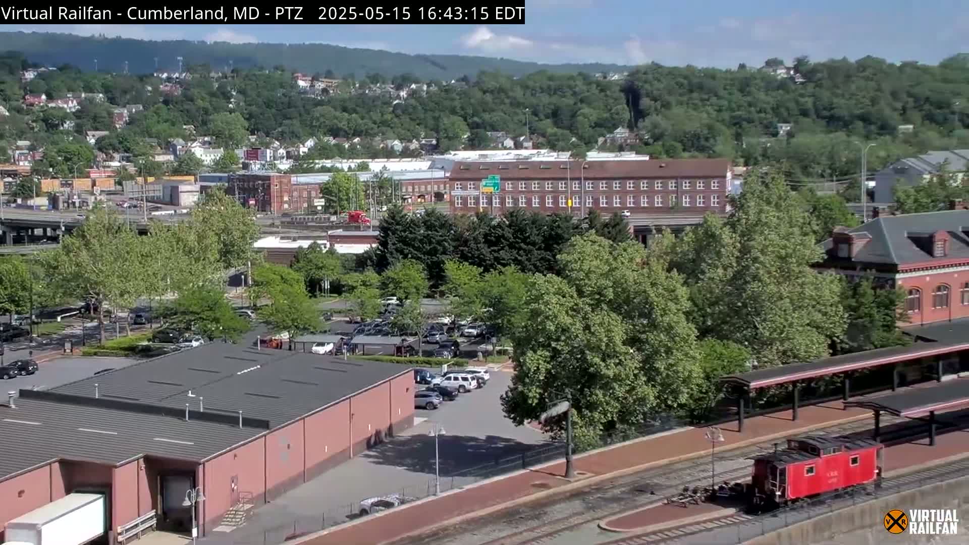 A mostly sunny view of a town with buildings, a parking lot, trees, and a red caboose next to a train platform.