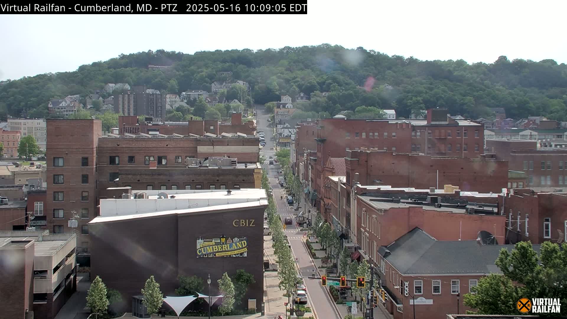 A mostly sunny view of a city street lined with brick buildings, nestled at the base of a tree-covered hill.