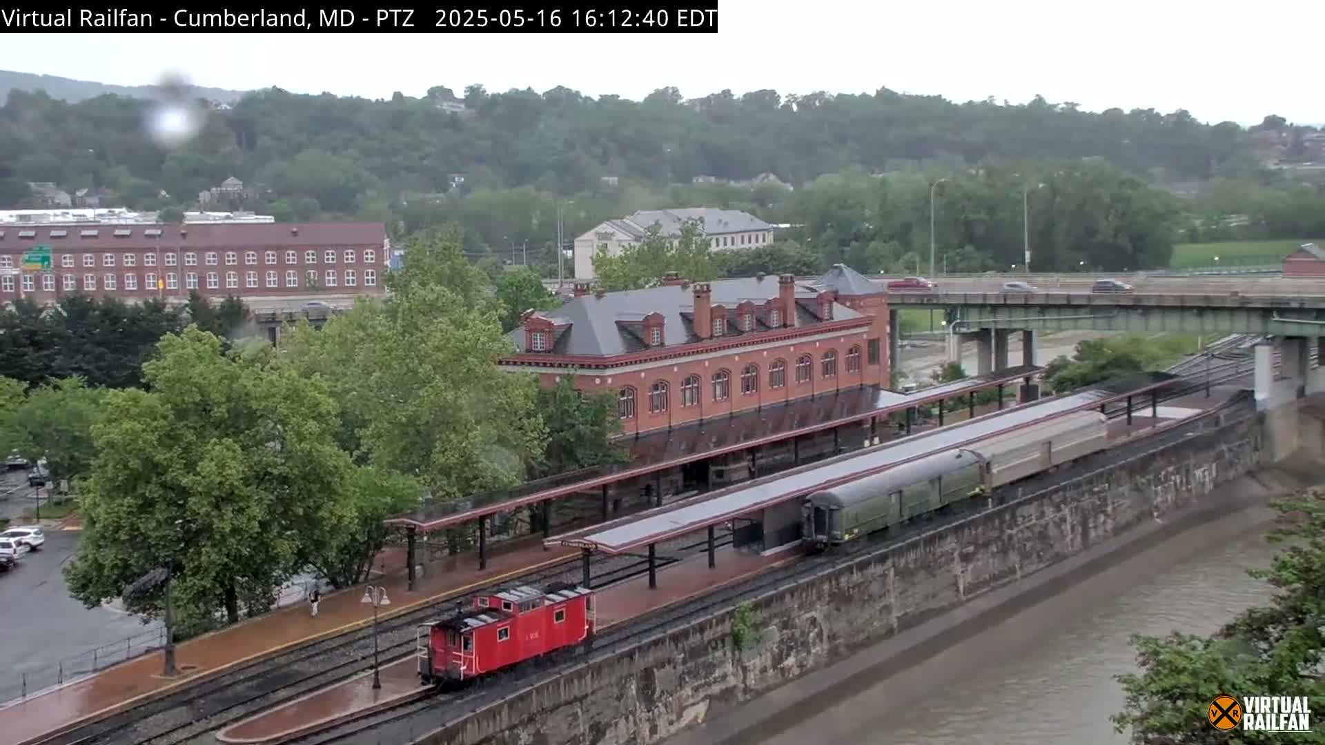 A red caboose and a long passenger train sit at a station beside a river under an overcast sky.