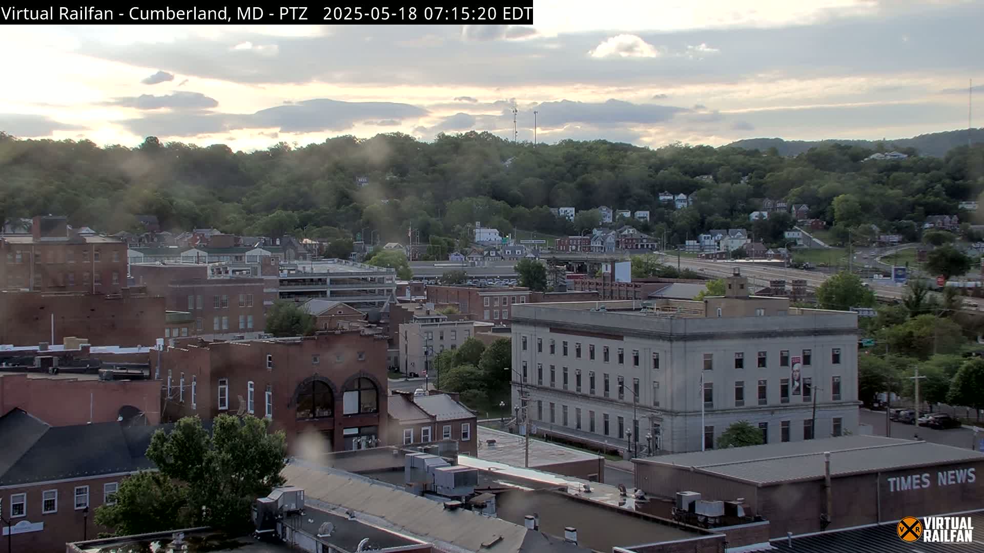 A slightly overcast view of a town nestled among green hills, showing brick buildings and a large light-colored building with many windows.