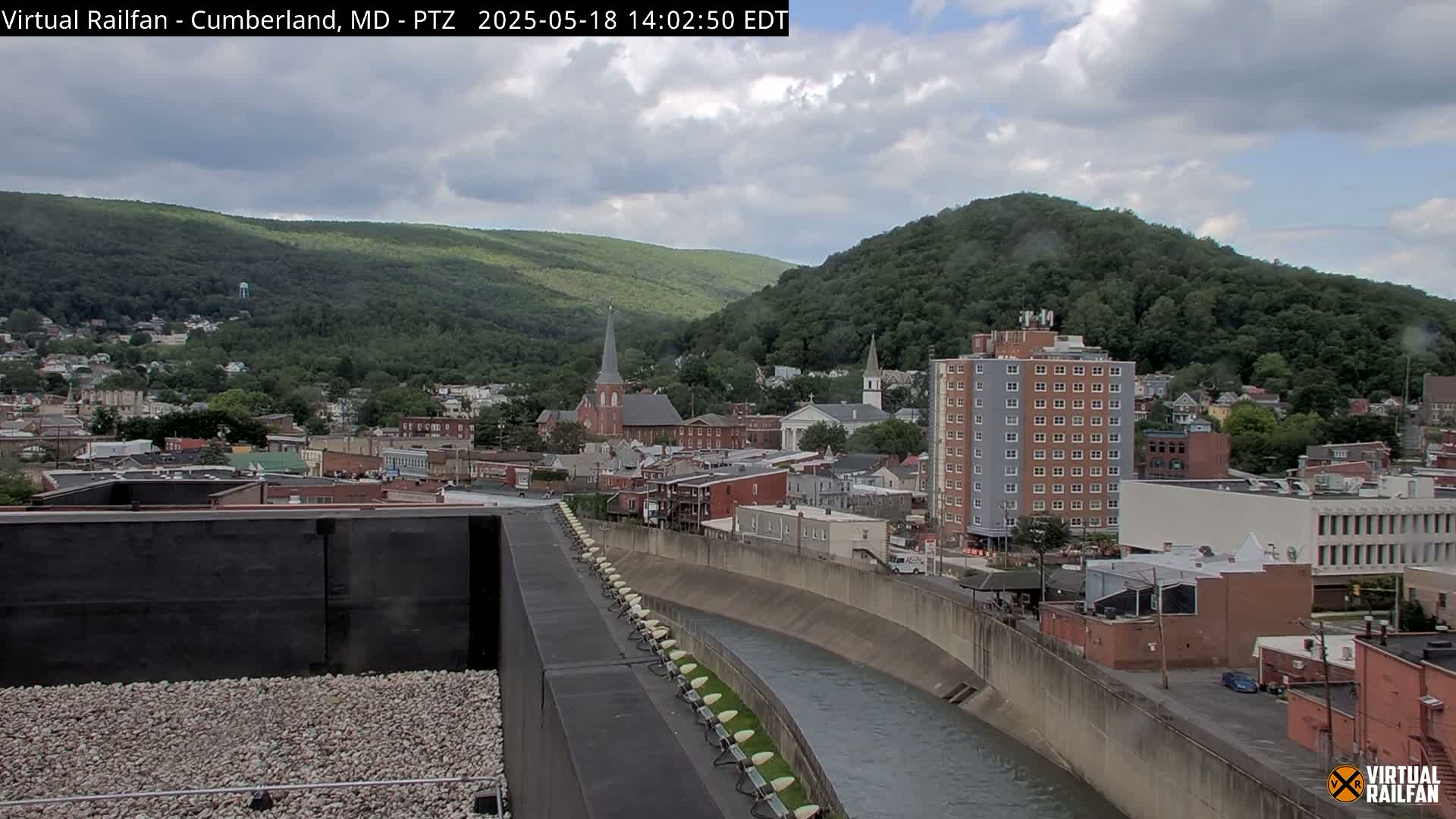 A partly cloudy day reveals a river curving through a town nestled in a valley, with hills covered in green trees in the background.
