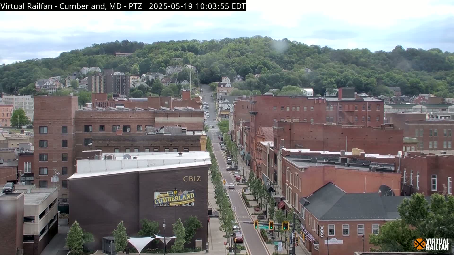 A long street lined with brick buildings in a town nestled below a green hill under a partly cloudy sky.