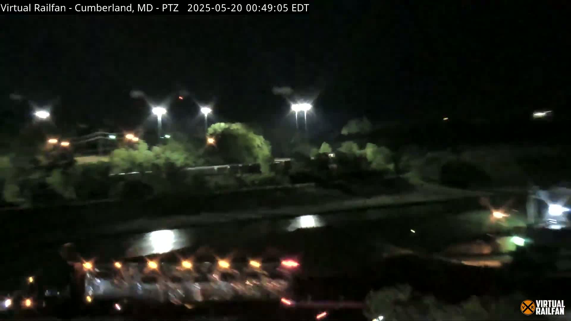 A nighttime view of a body of water under bright lights, with trees and what appears to be a train or a long line of vehicles along the bank.