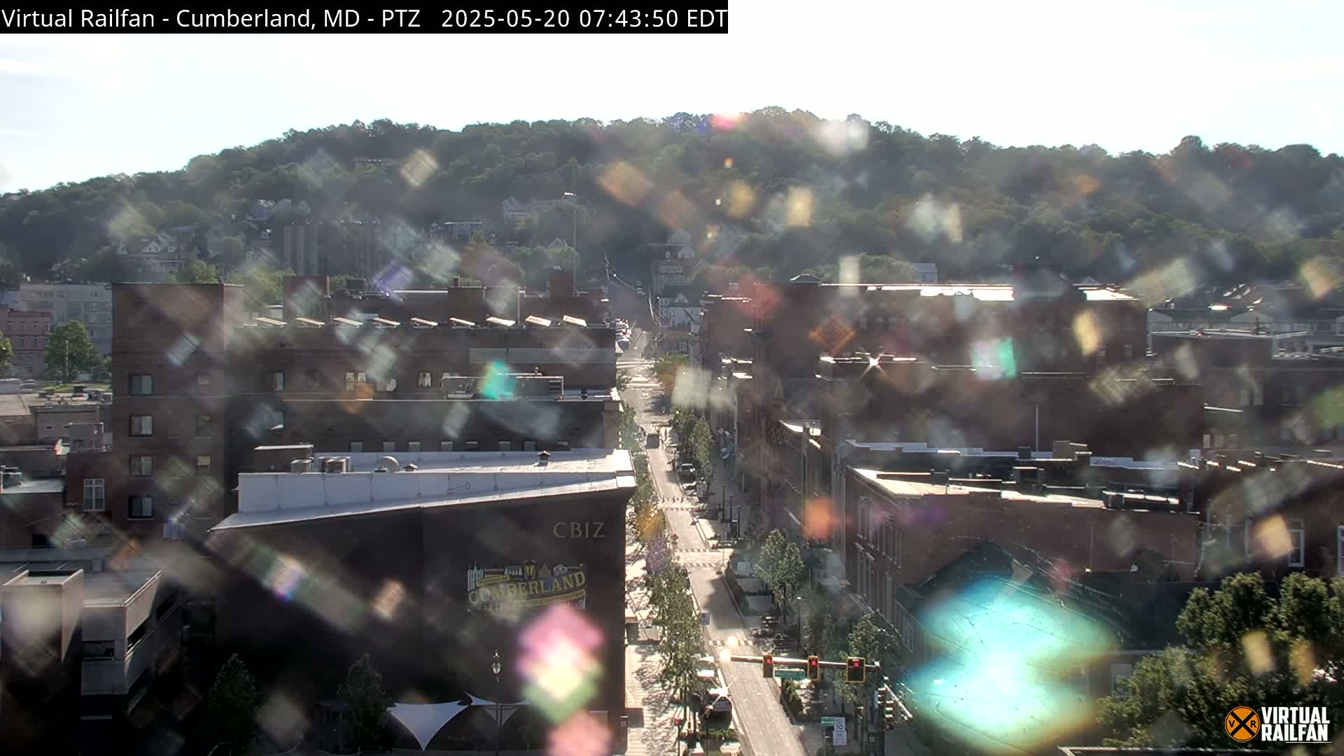 A sunny, high-angle view of a city street lined with brick buildings, nestled beneath a tree-covered hill.