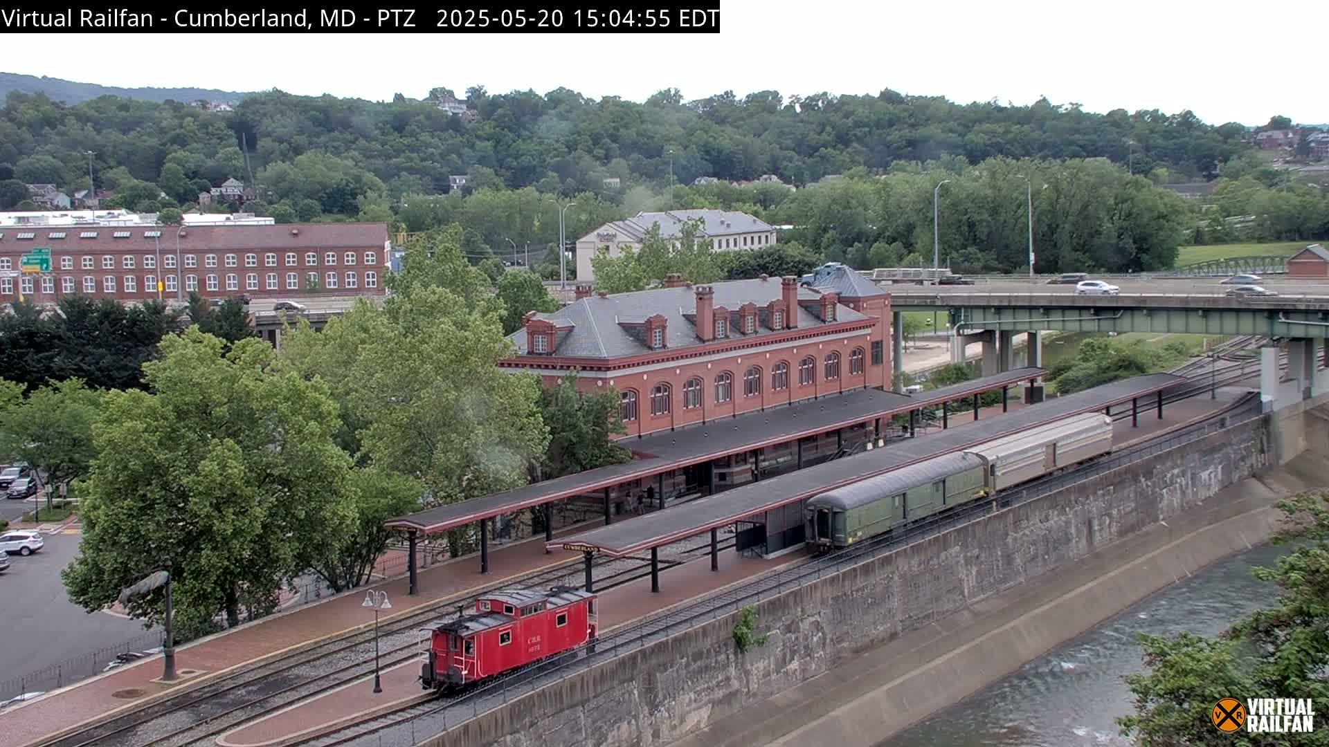 A red caboose and a long passenger train are parked at a brick train station next to a river under a partly cloudy sky.