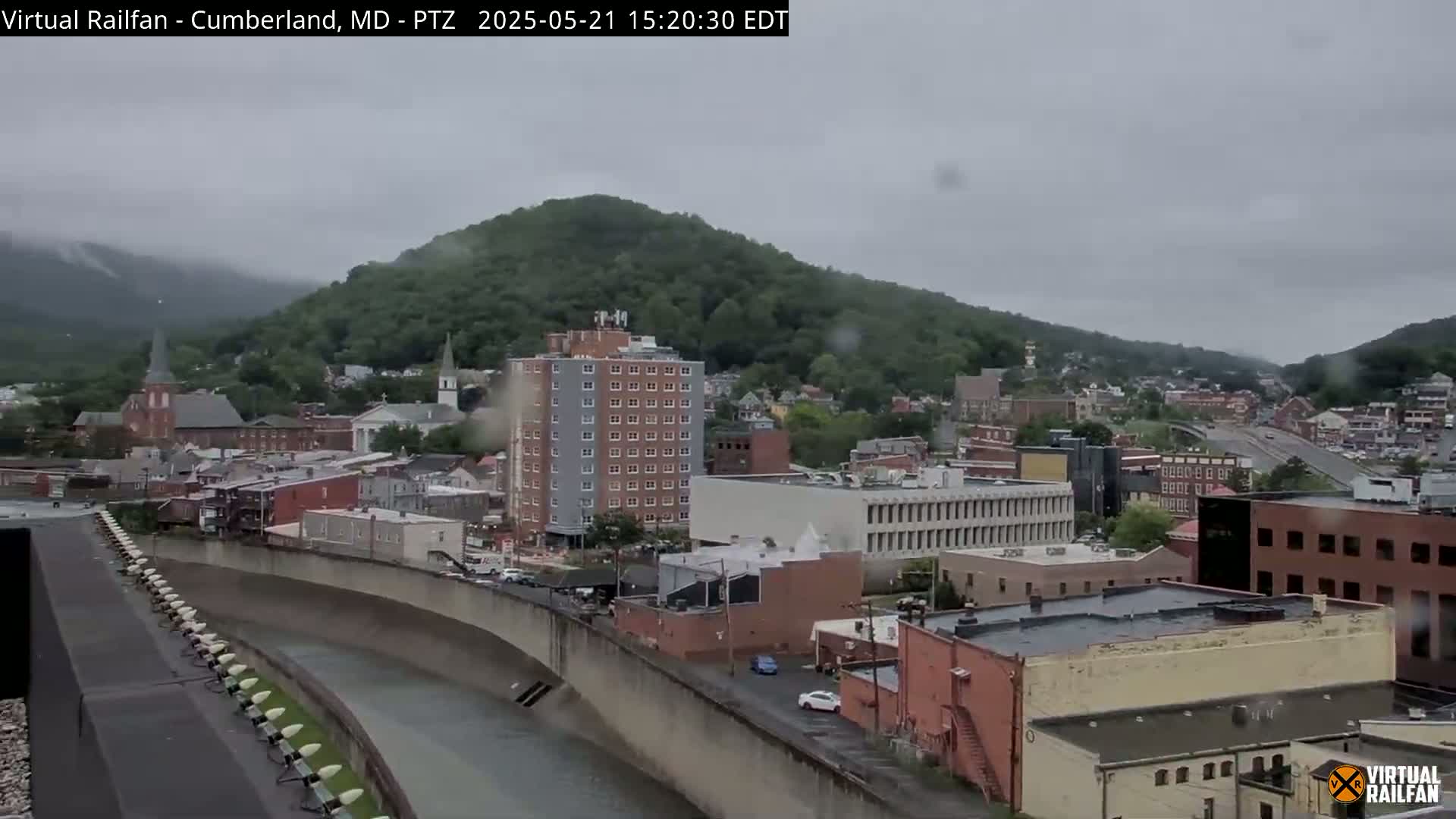 A rain-swept view of a town nestled in a valley, with a river flowing through it and hills covered in green trees in the background.