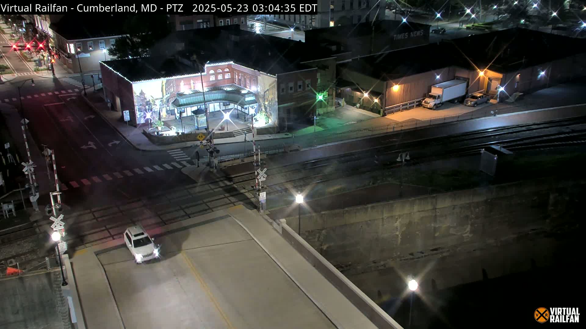 A nighttime, high-angle view of a city street intersection with railroad tracks, a white SUV, and several buildings.