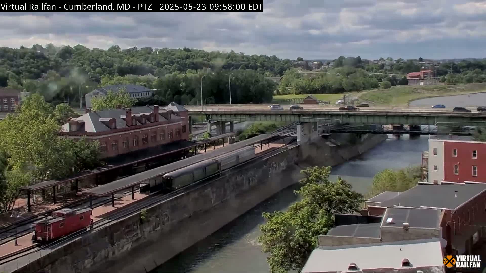 A train sits at a station beside a canal, under a partly cloudy sky, with cars traveling on a bridge overhead.