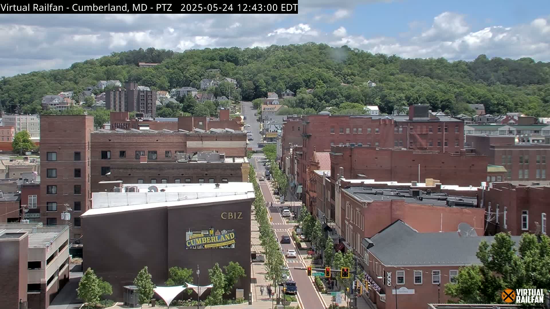 A partly cloudy day reveals a city street lined with brick buildings, leading toward a hill covered in lush green trees.