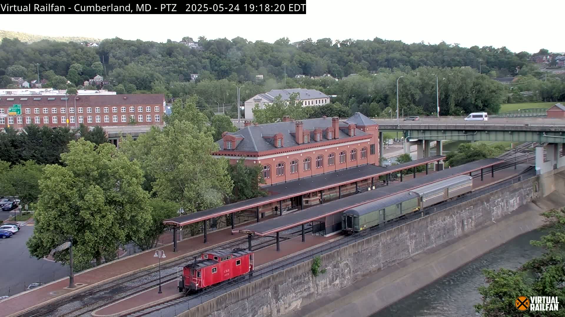 A red caboose and a long freight train are parked at a brick train station next to a river under a partly cloudy sky.