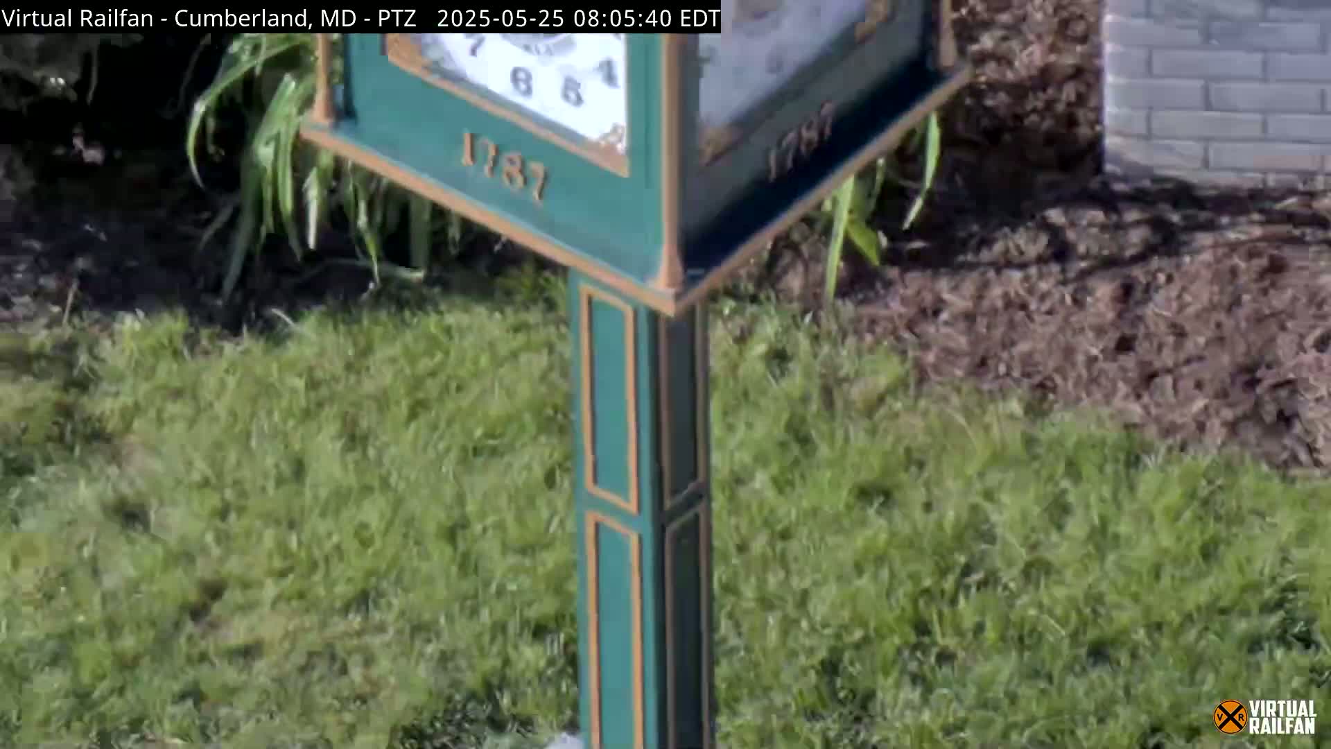 A green and gold clock post stands in a grassy area with wood chips at its base on a sunny day.