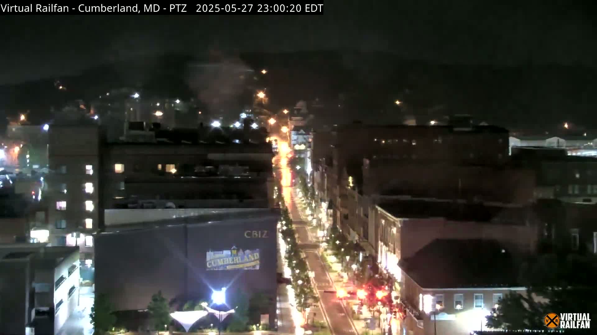 A nighttime, overhead view of a city street with buildings and streetlights illuminated under partly cloudy skies.