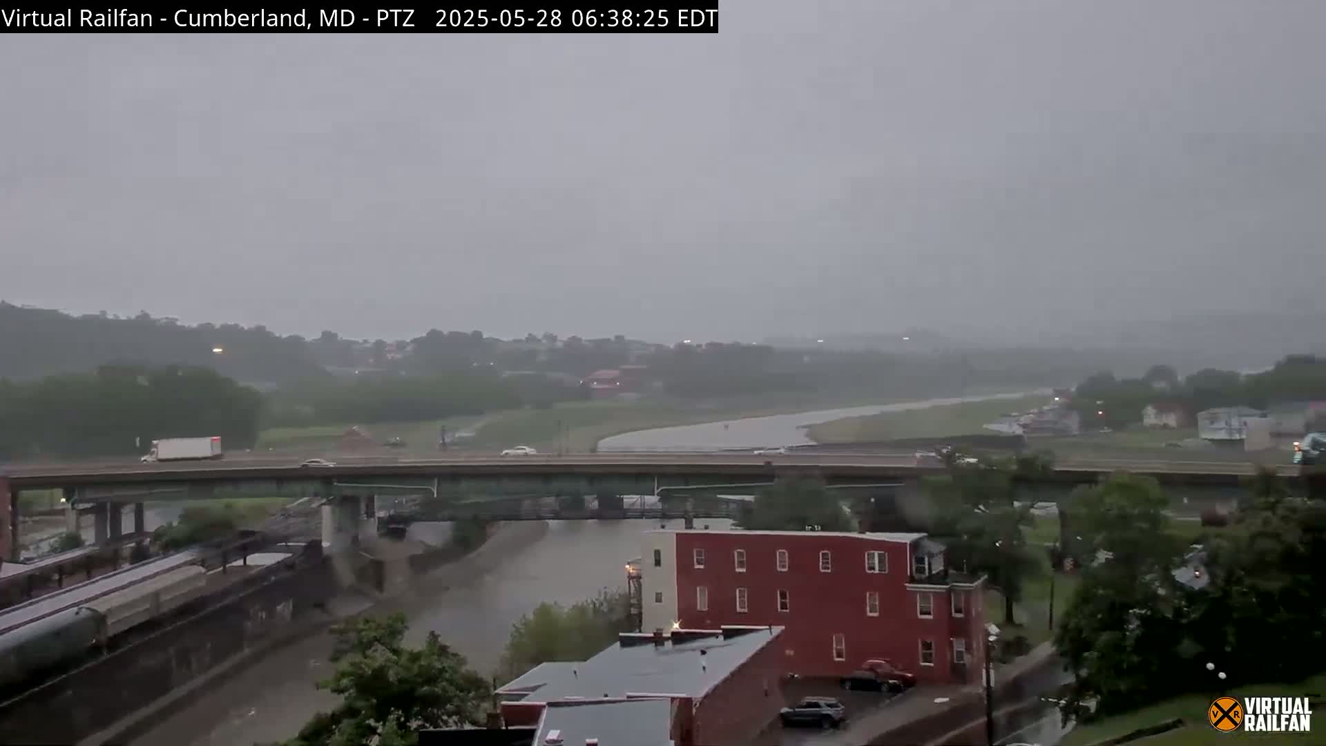 A rainy scene shows a river flowing under a bridge with a highway above it, cars driving on the highway, and buildings along the riverbank.