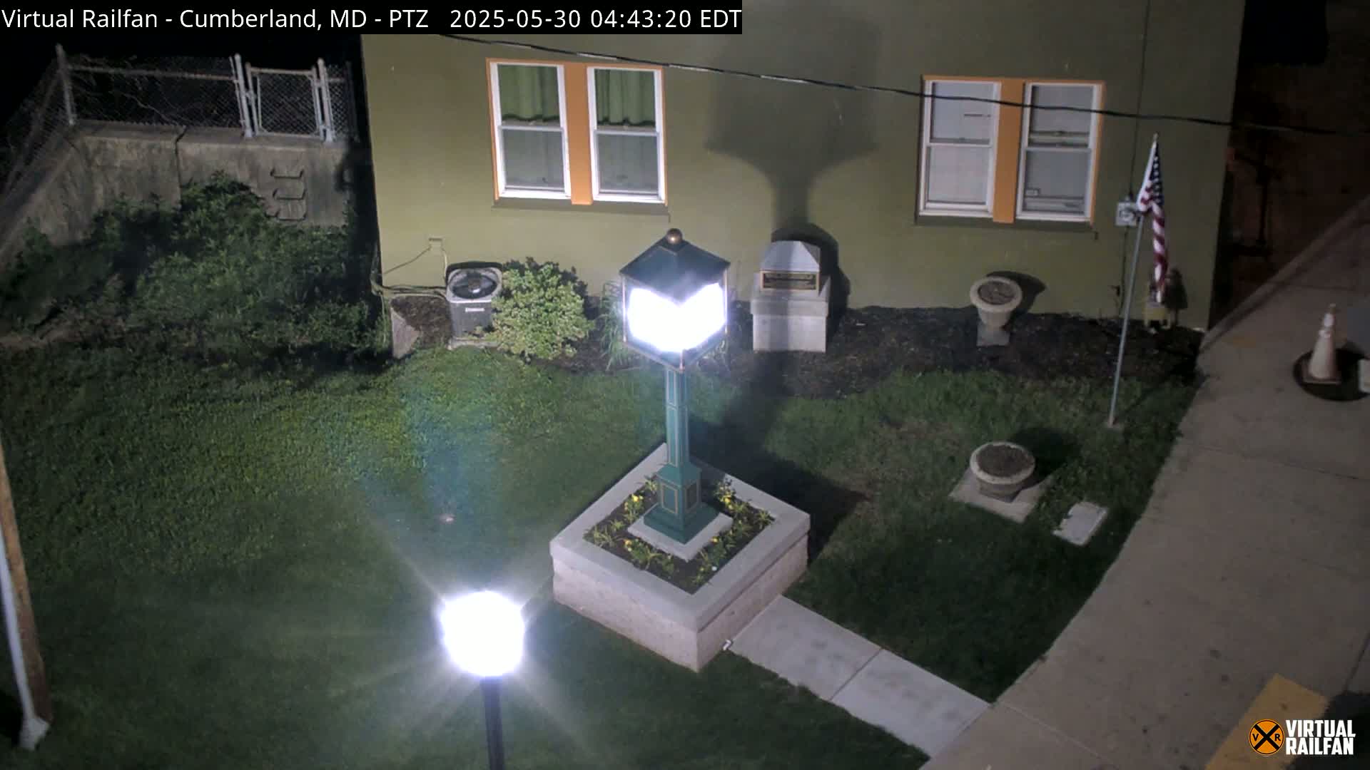 A brightly lit, green lamp post sits in a small, square planter surrounded by a grassy area next to a building at night.