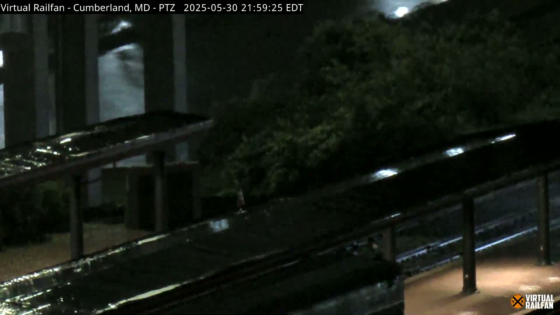 A nighttime, rain-soaked train platform is visible under a covered shelter, with rain reflecting off the wet surfaces.