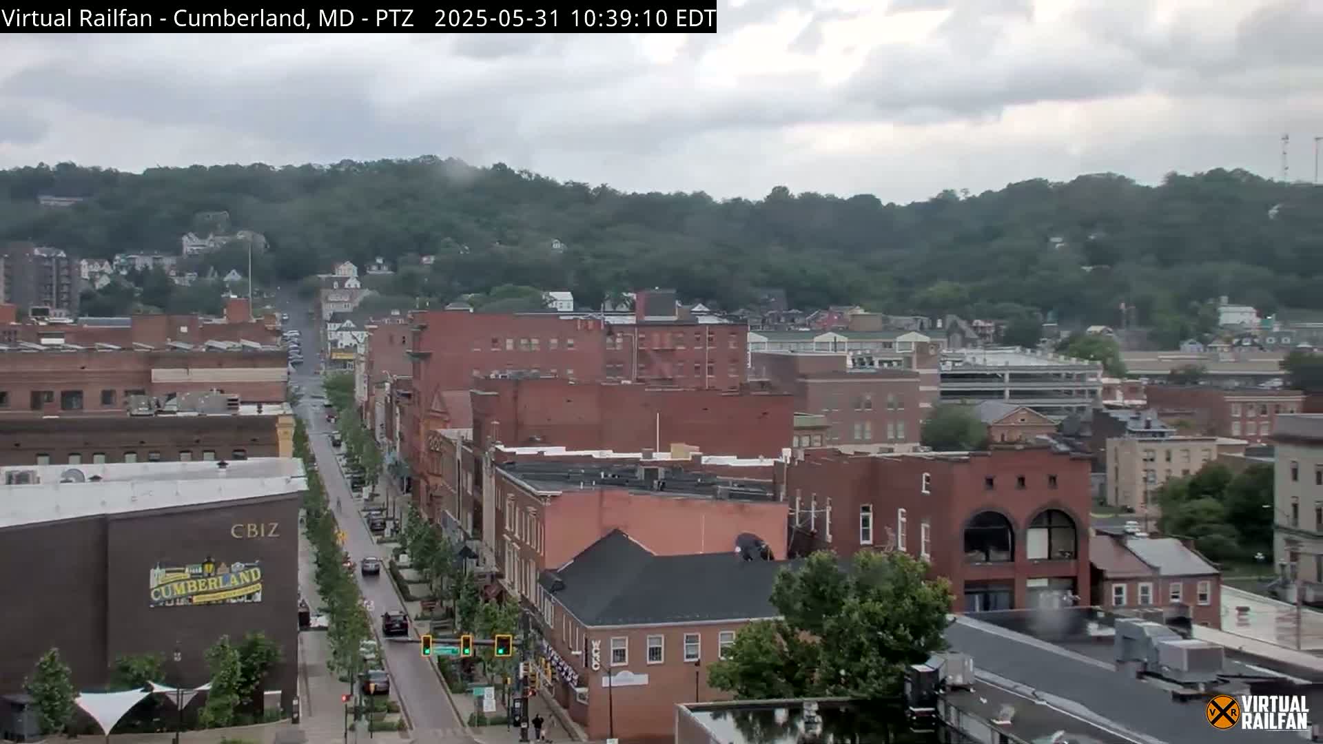 A city street scene on an overcast day shows several brick buildings, a tree-lined street with cars, and a hill covered in green trees in the background.
