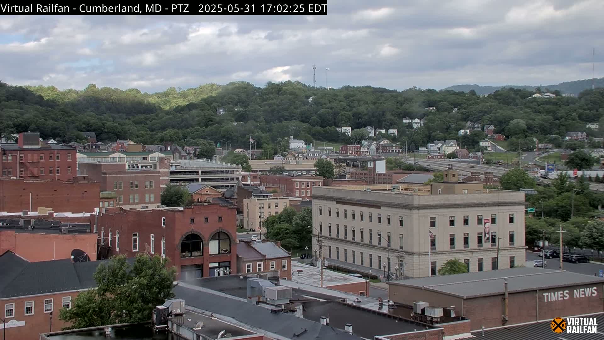 A partly cloudy day reveals a cityscape nestled against tree-covered hills, with numerous brick buildings and a prominent light-colored structure.