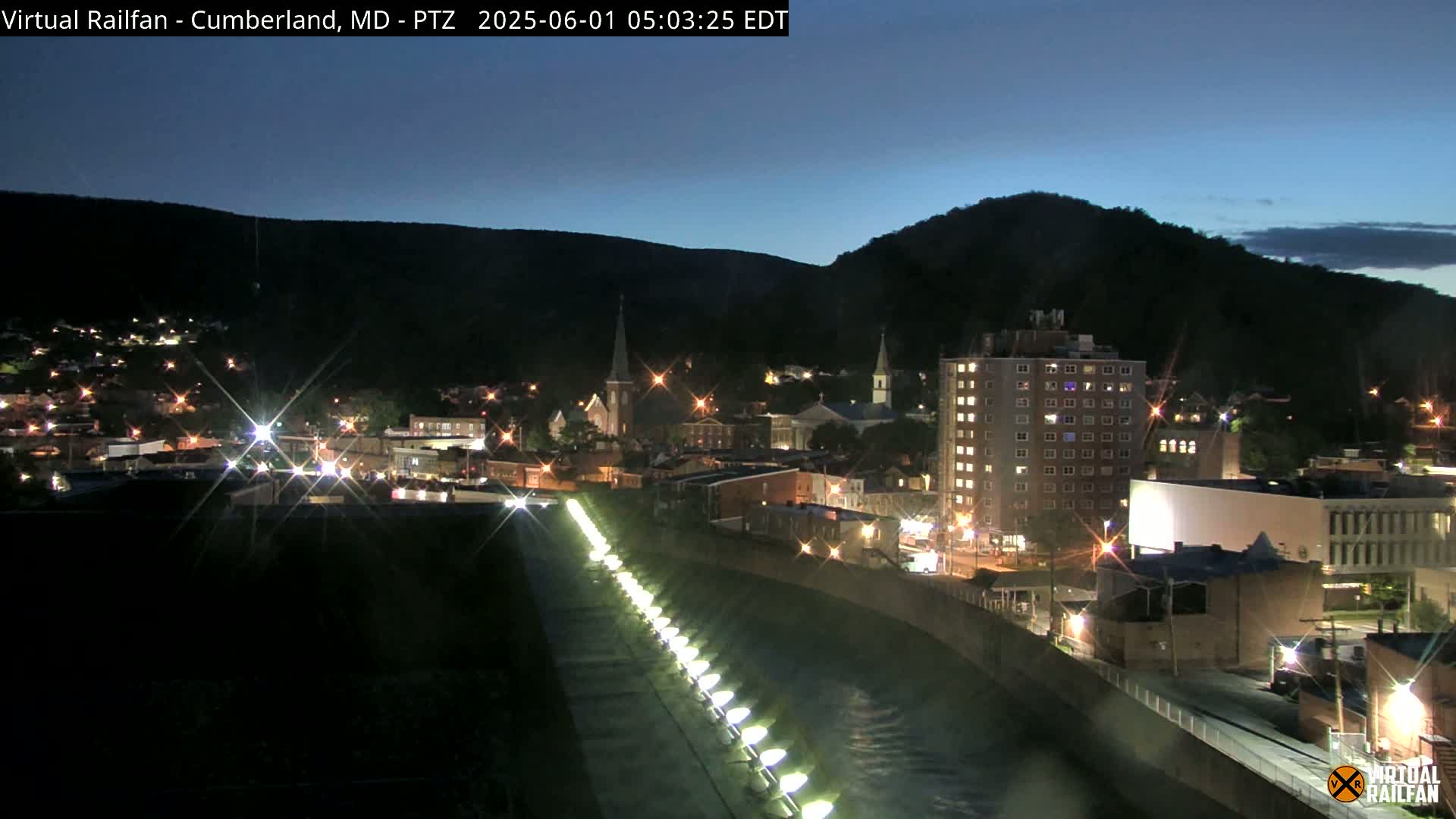 A nighttime view of a town nestled between mountains, illuminated by streetlights and building lights, with a river or canal in the foreground featuring a line of lights along its edge.